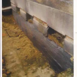 Odsłonięty fragment fundamentu z widocznymi otworami wentylacyjnymi, ziemia wokół. Ukazana struktura muru i detale wykonania. Prace ziemne w toku.