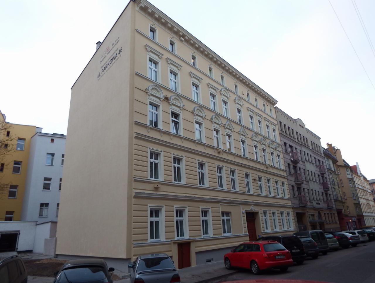 Odnowiona elewacja kamienicy w beżowych barwach, z widocznymi detalami architektonicznymi i zaparkowanymi autami. Budynek po remoncie, widok z ulicy.