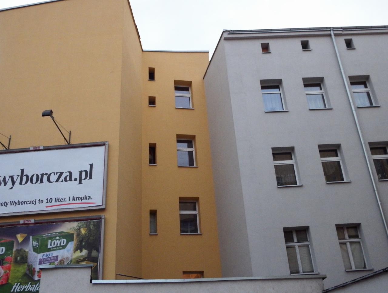 Dwa budynki o różnych kolorach elewacji: żółty z wąskimi oknami i szary z rurą spustową. Na pierwszym budynku reklama wyborcza.pl.