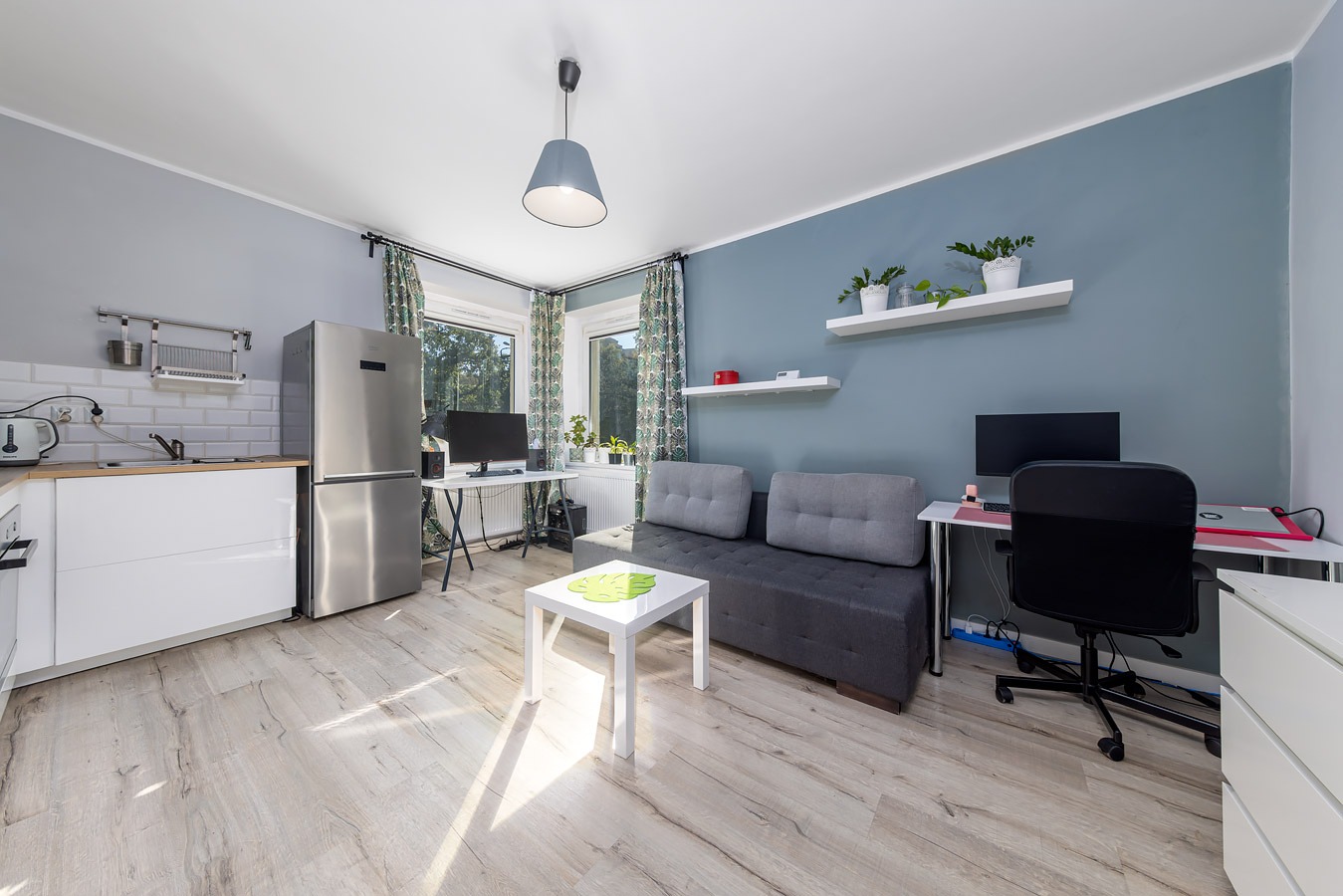 Jasne mieszkanie typu studio z aneksem kuchennym, szarą sofą z poduszkami, biurkiem z komputerem i drewnianą podłogą.