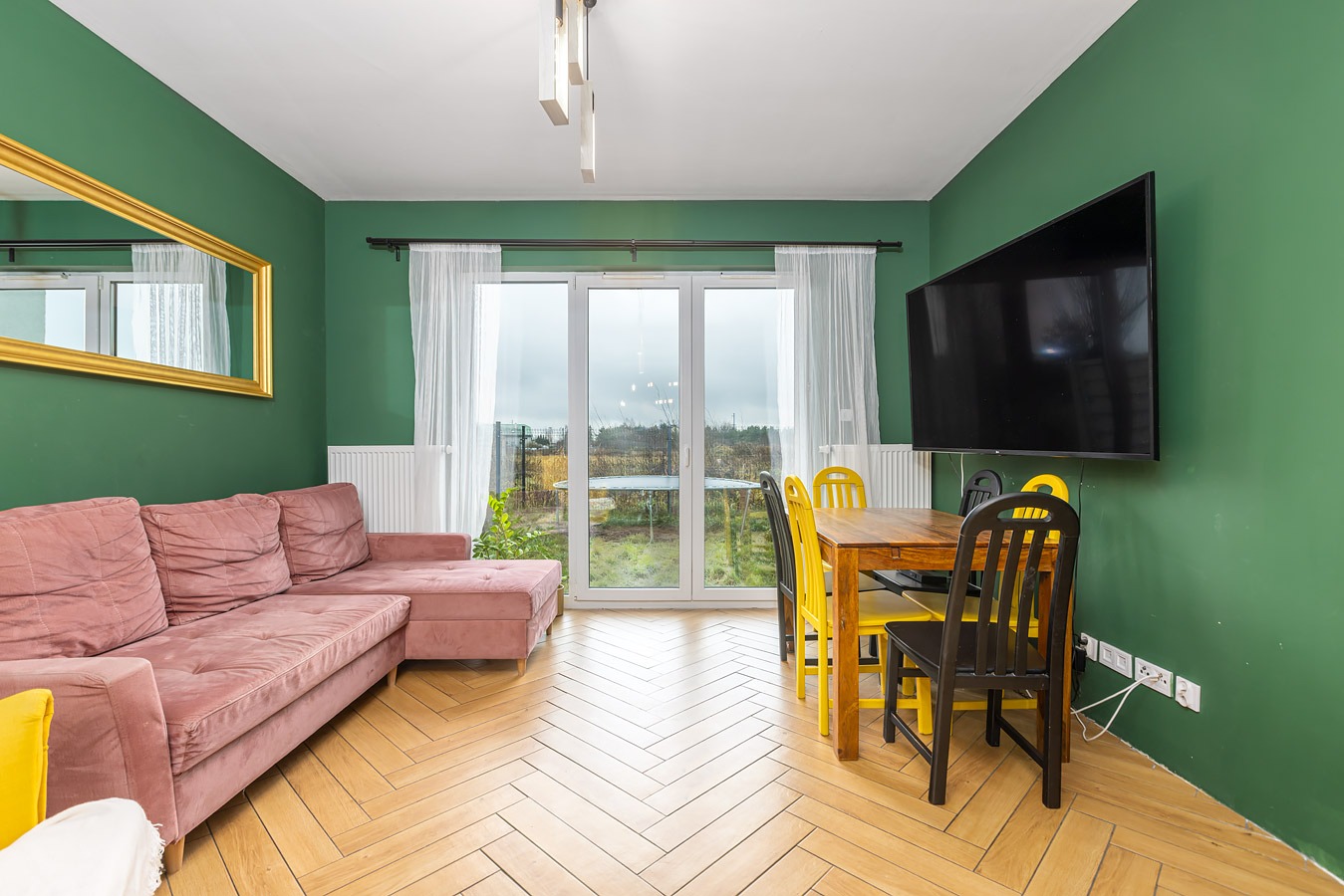 Wnętrze pokoju z zielonymi ścianami, parkietem w jodełkę, różową sofą, złotym lustrem, stołem z żółtymi i czarnymi krzesłami oraz dużym telewizorem na ścianie.