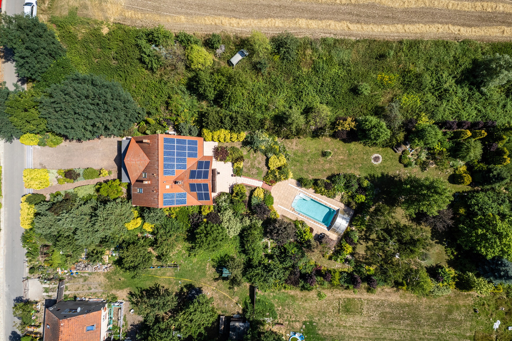 Widok z lotu ptaka na dom z czerwonym dachem i panelami słonecznymi, otoczony bujną zielenią, basenem z zadaszeniem i zadbanym ogrodem.