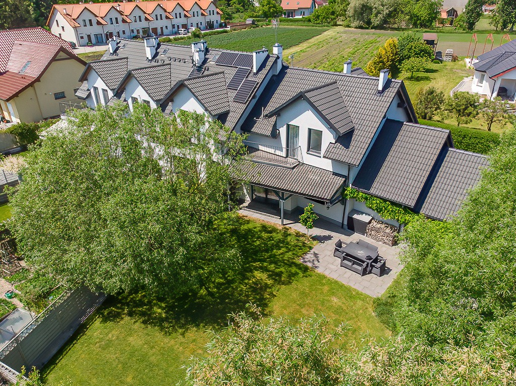 Widok z góry na szeregowy dom z ciemnoszarą dachówką, panelami słonecznymi i zielonym ogrodem z patio oraz zestawem mebli ogrodowych.