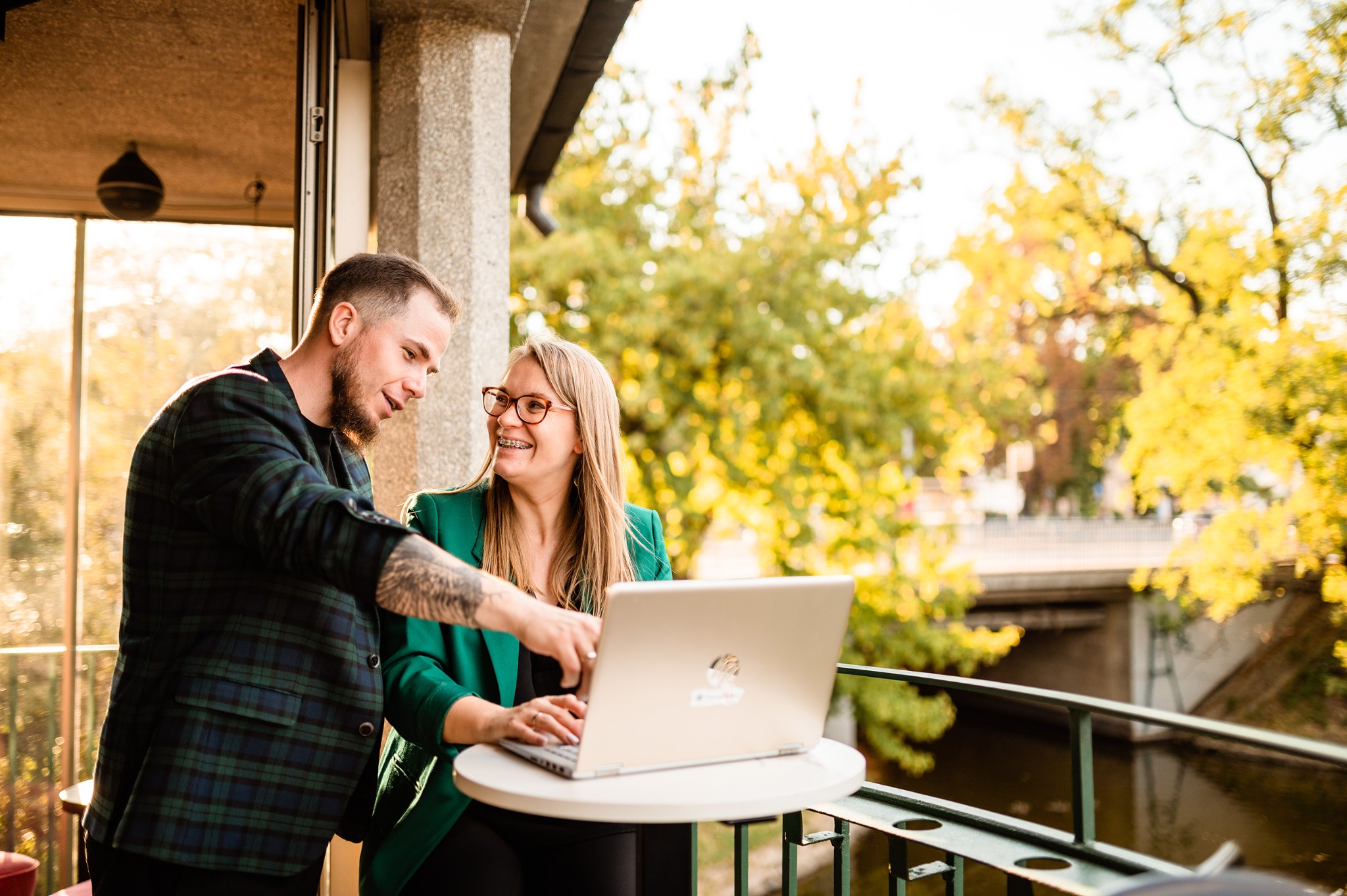 Dwie osoby, mężczyzna z tatuażem na ramieniu i kobieta w okularach i zielonej marynarce, pracują razem przy laptopie na balkonie z widokiem na drzewa i rzekę.