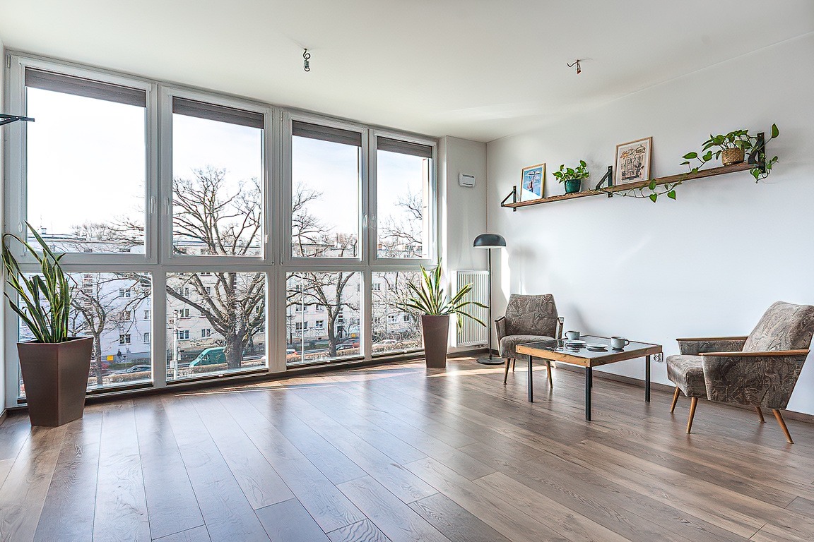 Jasne wnętrze salonu z dużym oknem, drewnianą podłogą, dwoma fotelami w stylu vintage, stolikiem kawowym z czarnymi nogami i roślinami doniczkowymi.