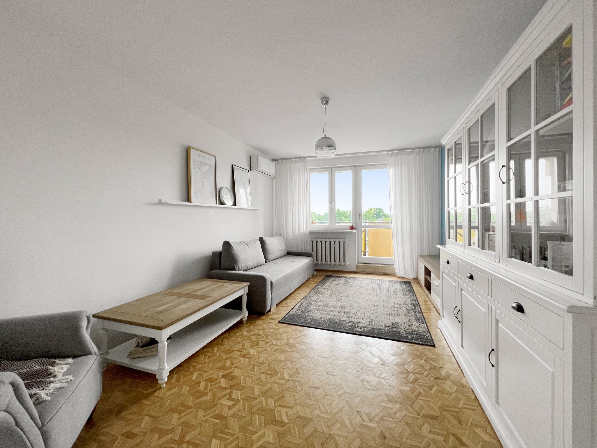 Jasne wnętrze pokoju dziennego z drewnianą podłogą w jodełkę, szarą sofą, białym kredensem z przeszkleniami i balkonem z widokiem na zieleń.