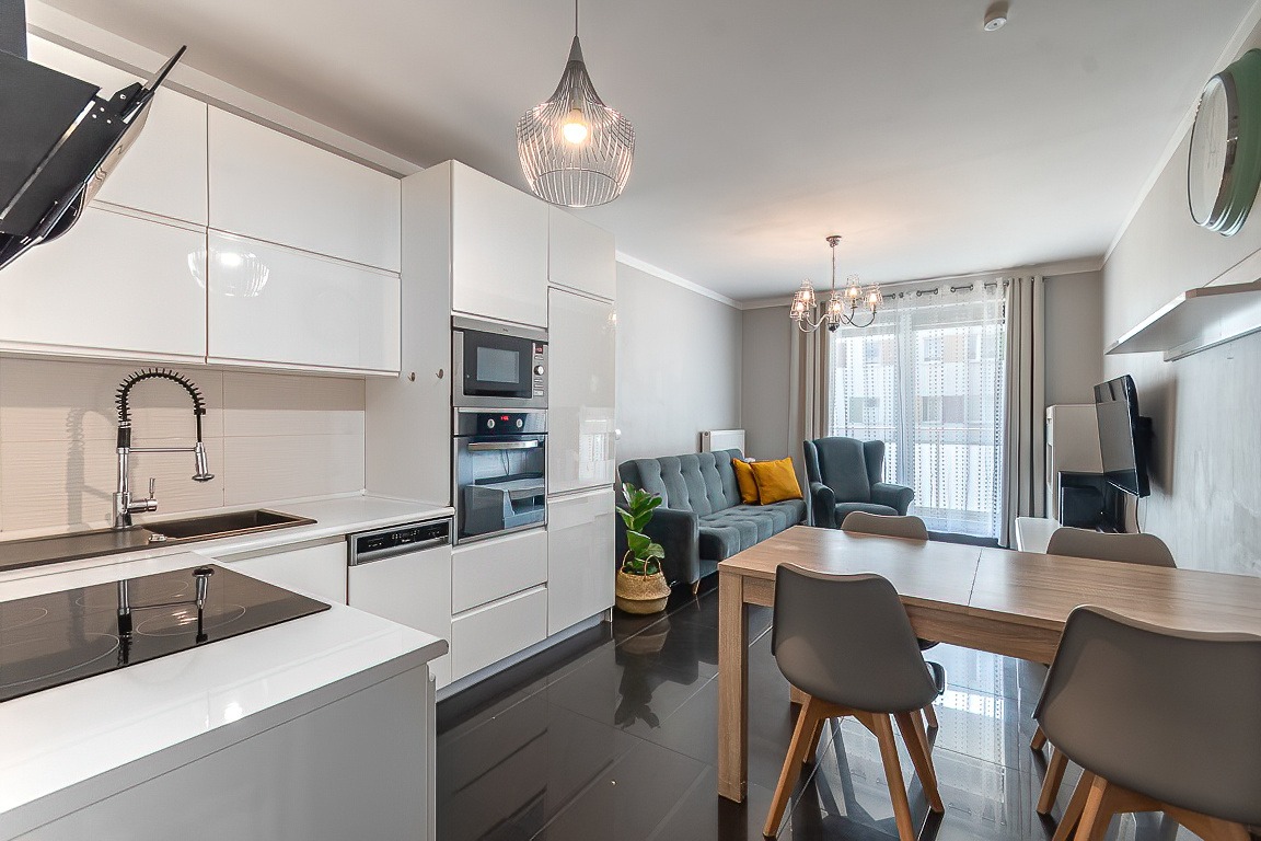 Jasne, nowoczesne wnętrze mieszkania z otwartą kuchnią, szarym narożnikiem z żółtymi poduszkami i drewnianym stołem z szarymi krzesłami.