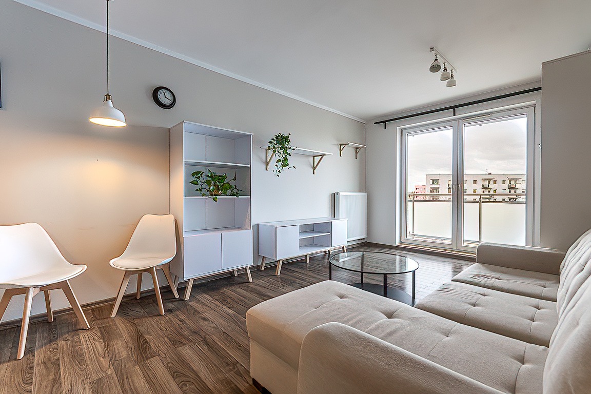 Jasne, przestronne wnętrze salonu z beżową sofą modułową, okrągłym szklanym stolikiem kawowym, białymi meblami i widokiem na budynek mieszkalny z balkonami za dużym oknem balkonowym.