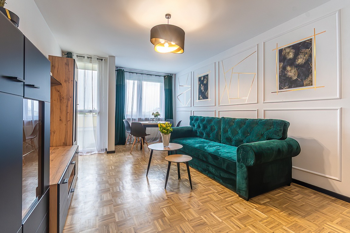 Wnętrze salonu z parkietem, szmaragdową sofą Chesterfield, stolikiem kawowym z bukietem tulipanów, ścianą z dekoracyjnymi listwami i obrazami w złotych ramach, oraz jadalnią w tle.