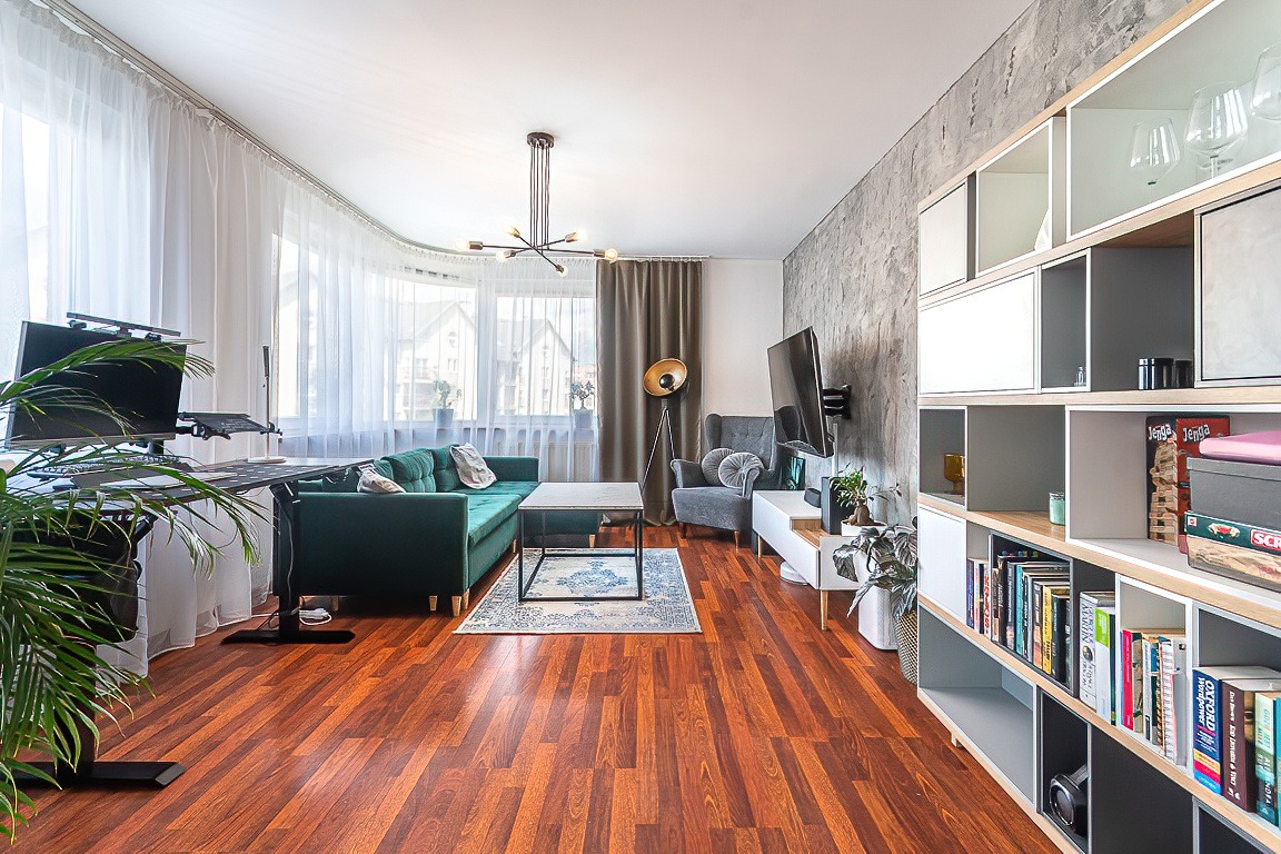 Jasne wnętrze salonu z zieloną sofą, szarym fotelem, biurkiem z komputerem i regałem z książkami, oświetlone przez duże okna z białymi zasłonami.