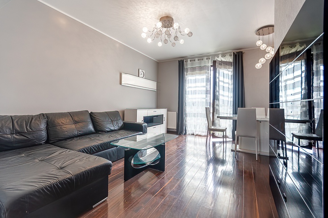 Jasne wnętrze salonu z czarną skórzaną sofą narożną, szklanym stolikiem kawowym, białymi meblami pod telewizorem, stołem z krzesłami i dekoracyjnymi lampami sufitowymi.