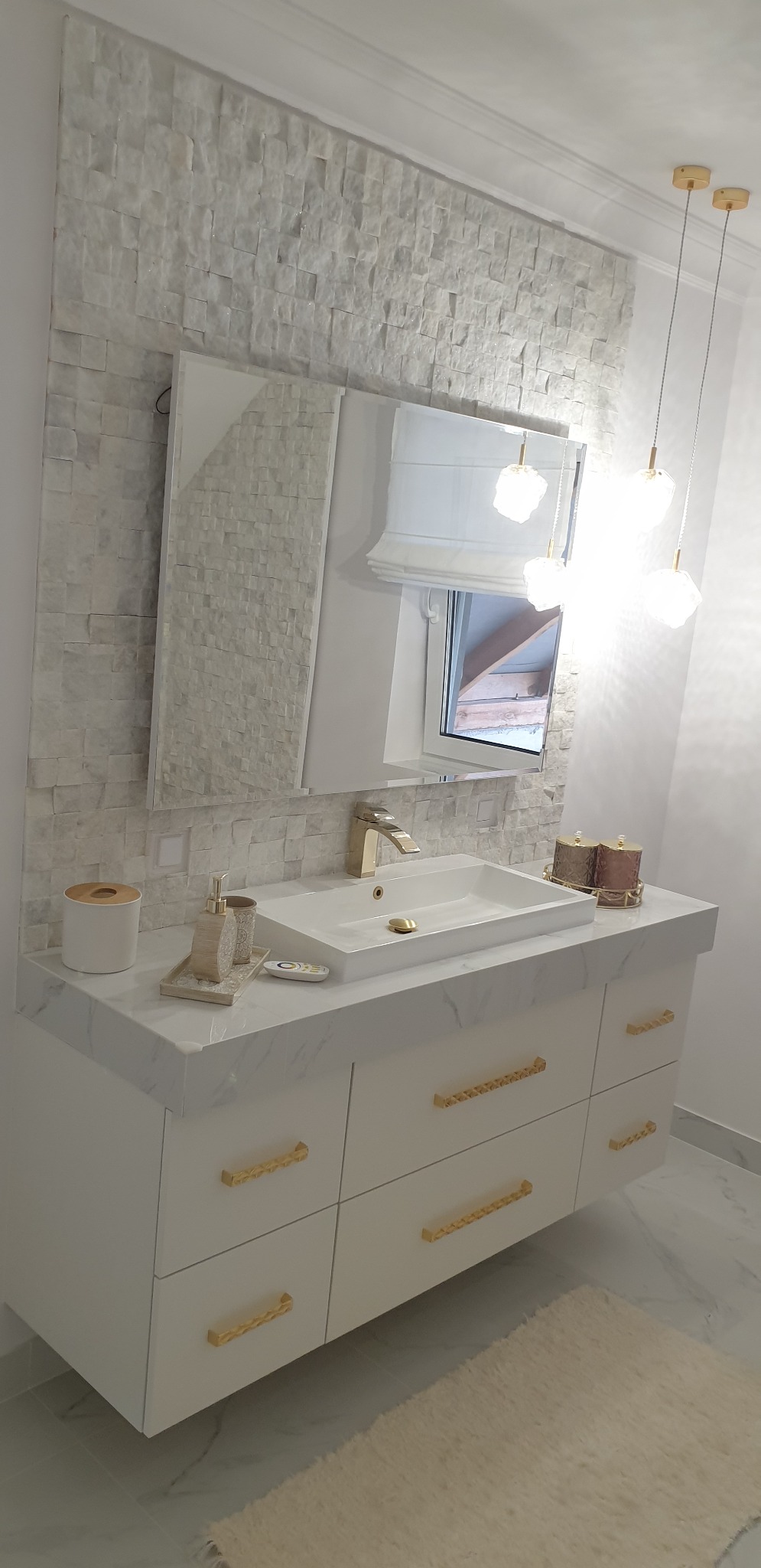 Nowoczesna łazienka z białymi szafkami z marmurowym blatem, złotymi uchwytami, prostokątną umywalką, złotą armaturą, lustrem, oświetleniem wiszącym i ścianą wyłożoną dekoracyjną mozaiką.