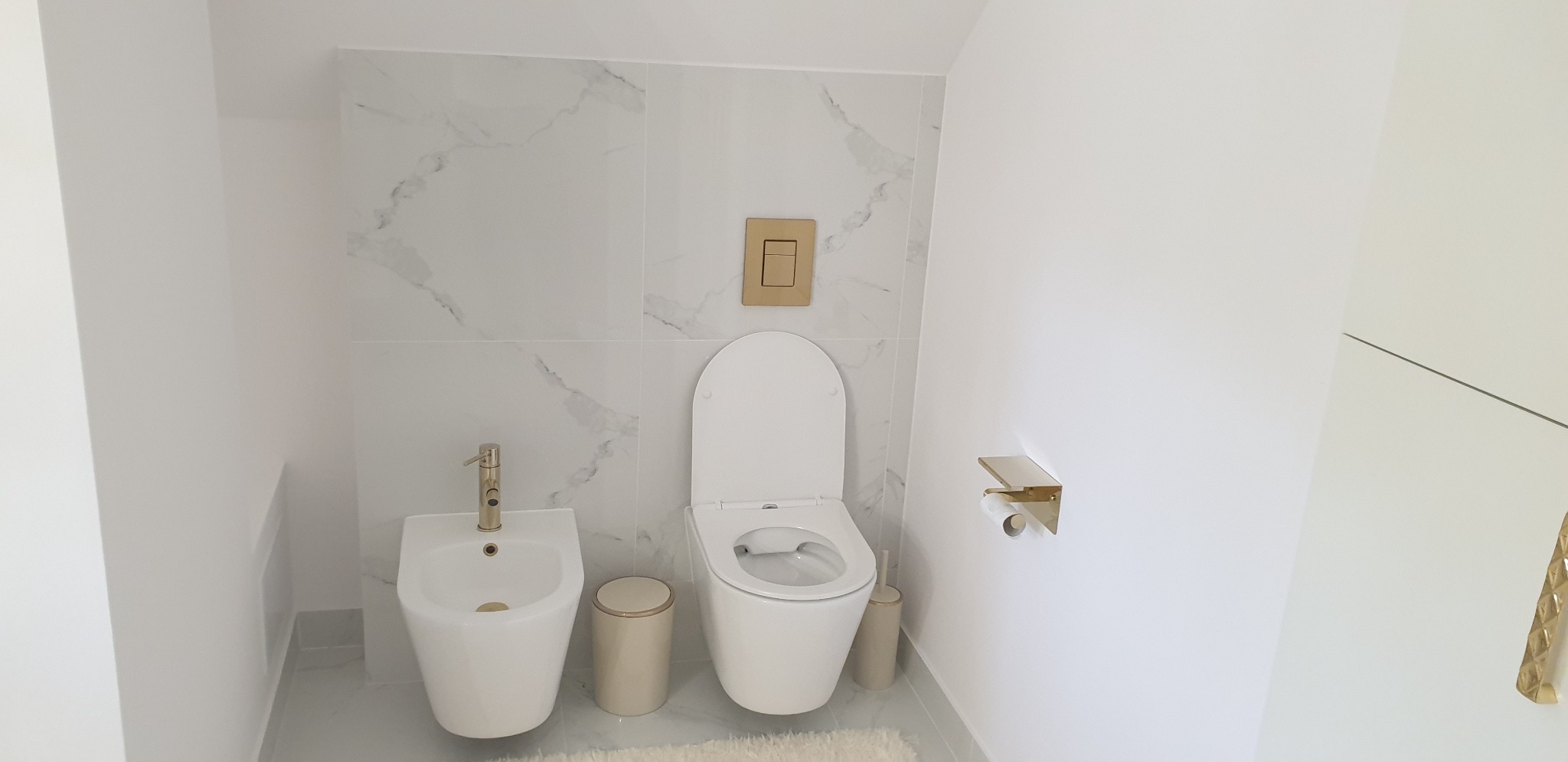 Biała łazienka z marmurowymi płytkami, złotymi akcentami w postaci armatury, przycisku spłuczki i uchwytu na papier toaletowy, z bidetem i podwieszaną toaletą.