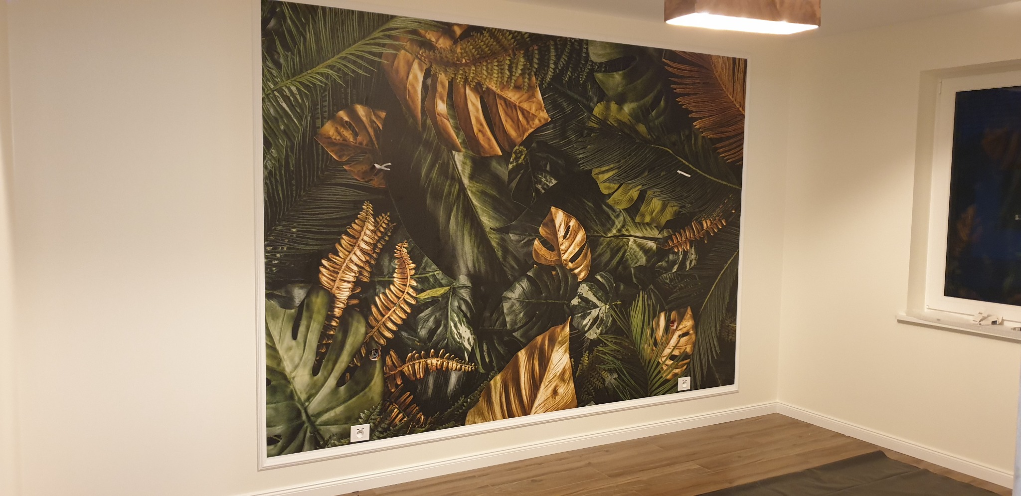 Ściana w pokoju z dużą fototapetą przedstawiającą tropikalne liście w odcieniach zieleni i złota, oświetlona górnym światłem, widoczny fragment podłogi z paneli i okno.