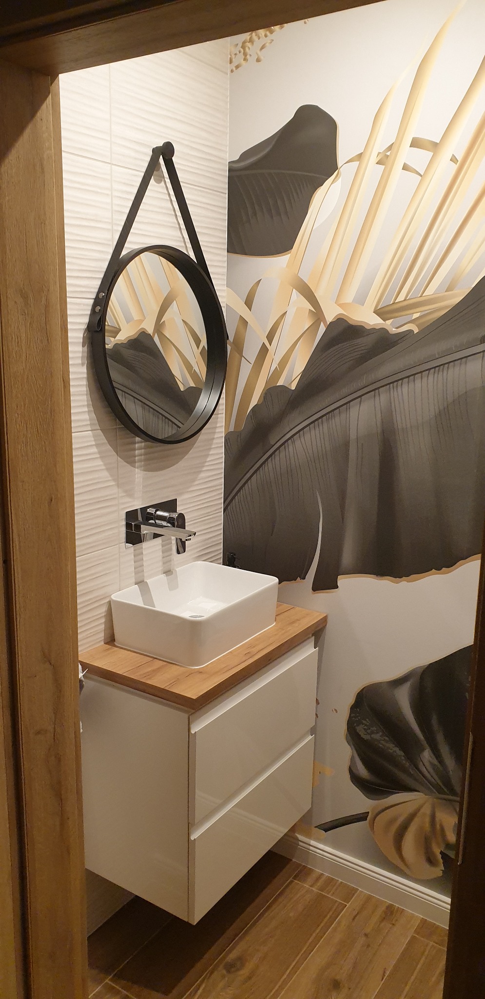 Nowoczesna łazienka z białą umywalką nablatową, okrągłym lustrem na pasku i tapetą w motywy liści bananowca, drewnopodobne płytki na podłodze.