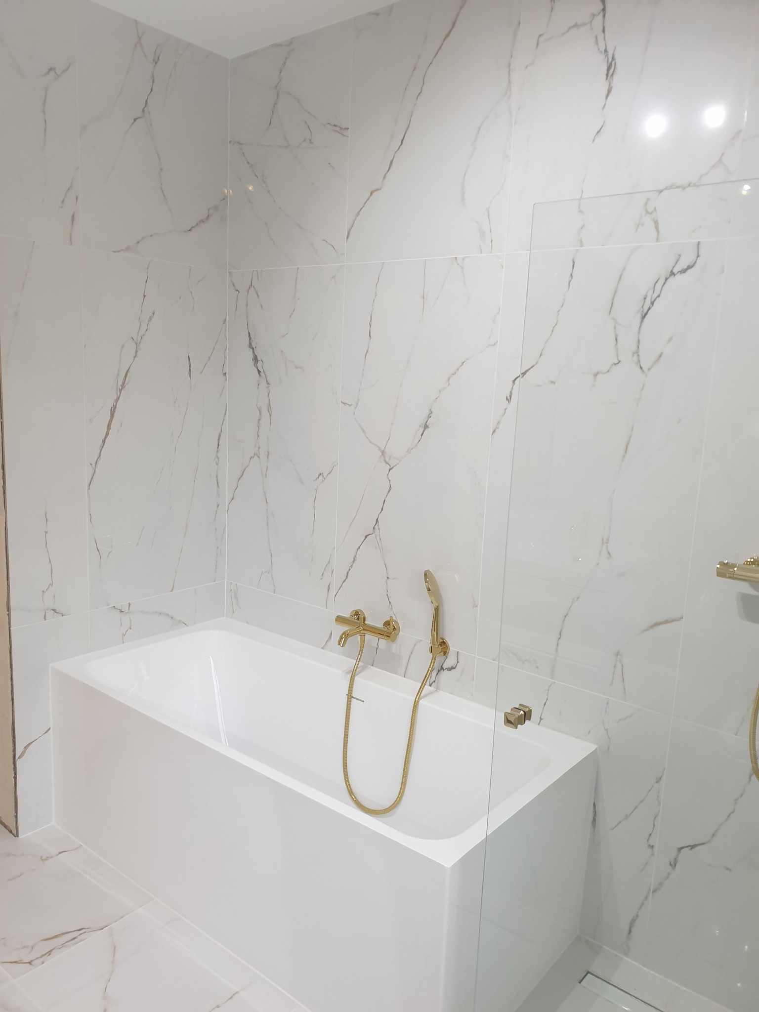 Nowoczesna łazienka z białą wanną obudowaną płytkami imitującymi marmur i złotą armaturą.