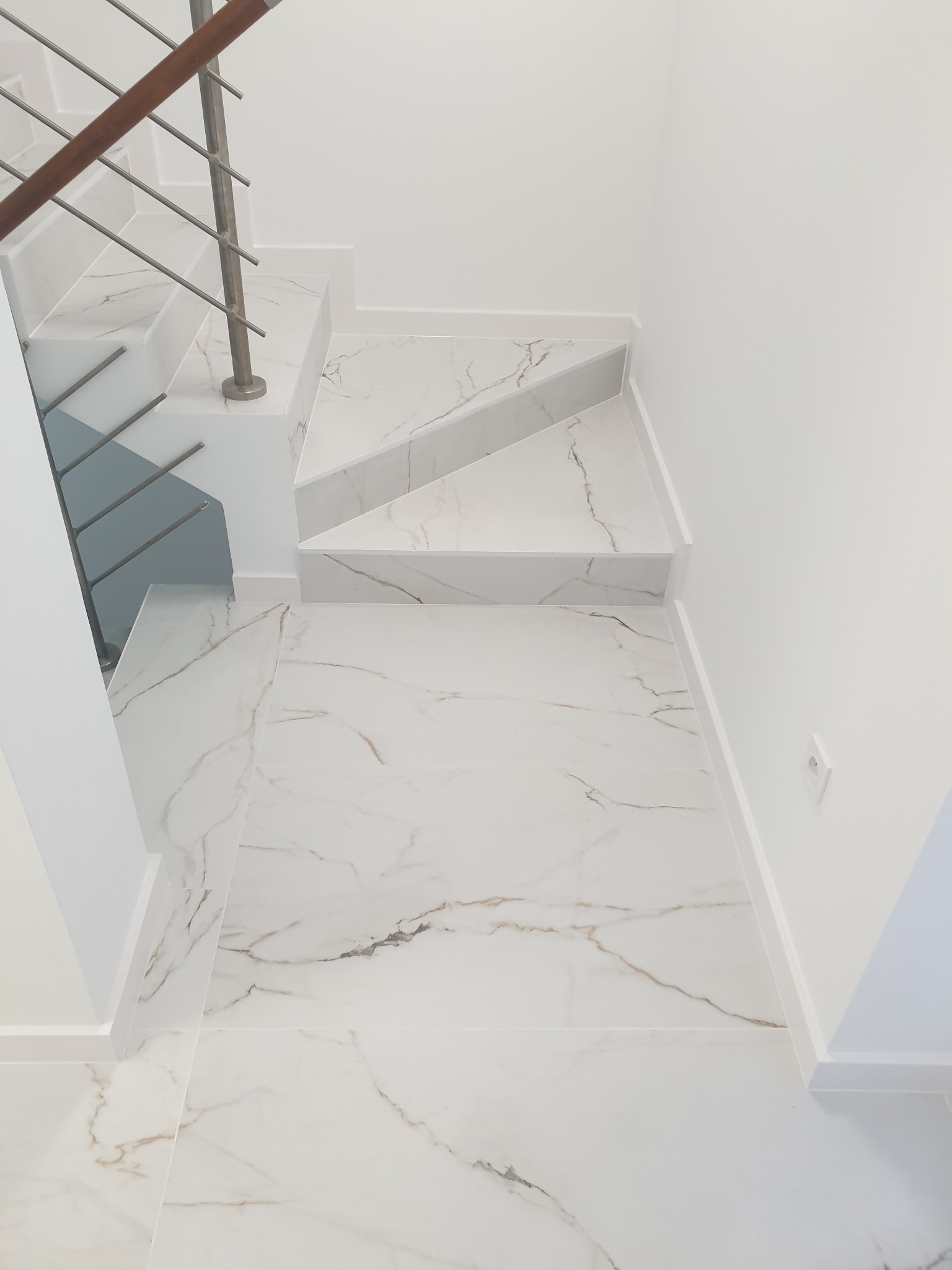 Białe marmurowe schody z metalową balustradą i drewnianym poręczą, widok z góry na wykończoną podłogę.