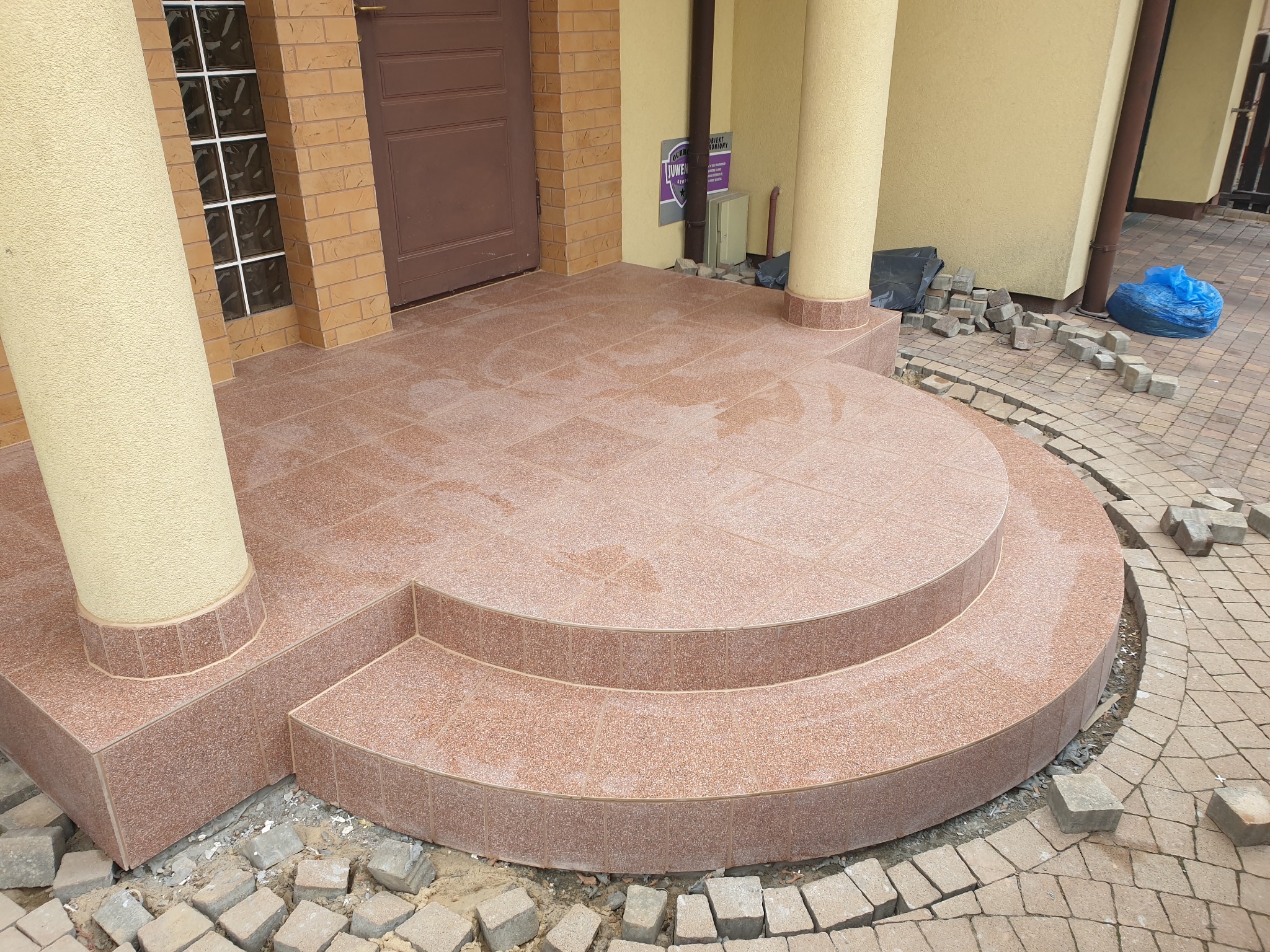 Wykończony taras z dwoma półokrągłymi stopniami, wyłożony płytkami w odcieniach brązu, z kolumnami i kostką brukową w trakcie układania na zewnątrz.