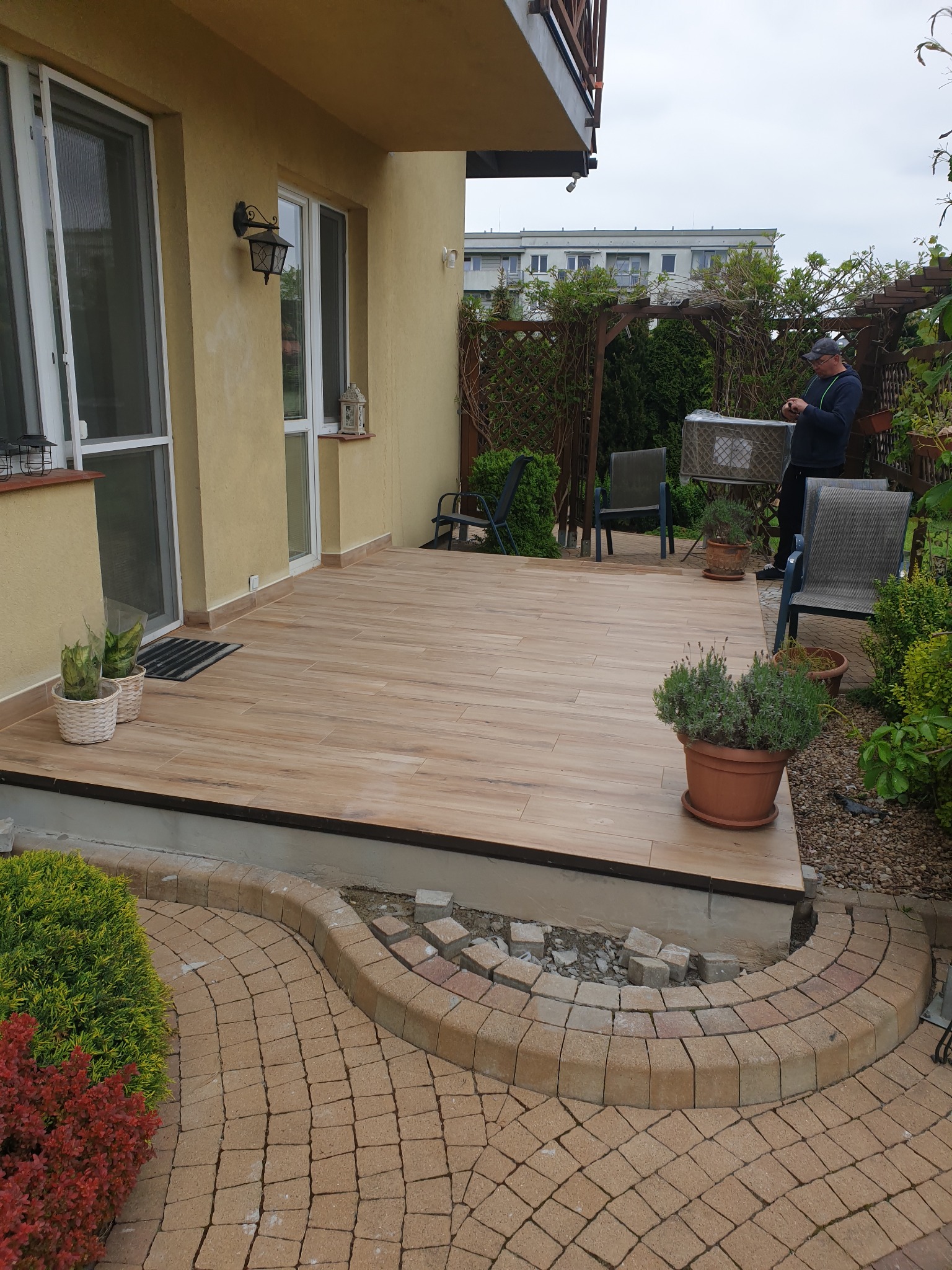 Wyłożony drewnopodobnymi płytkami taras przydomowy z widocznym fragmentem obrzeża z kostki, częściowo jeszcze nieukończonego, oraz osobą w tle.