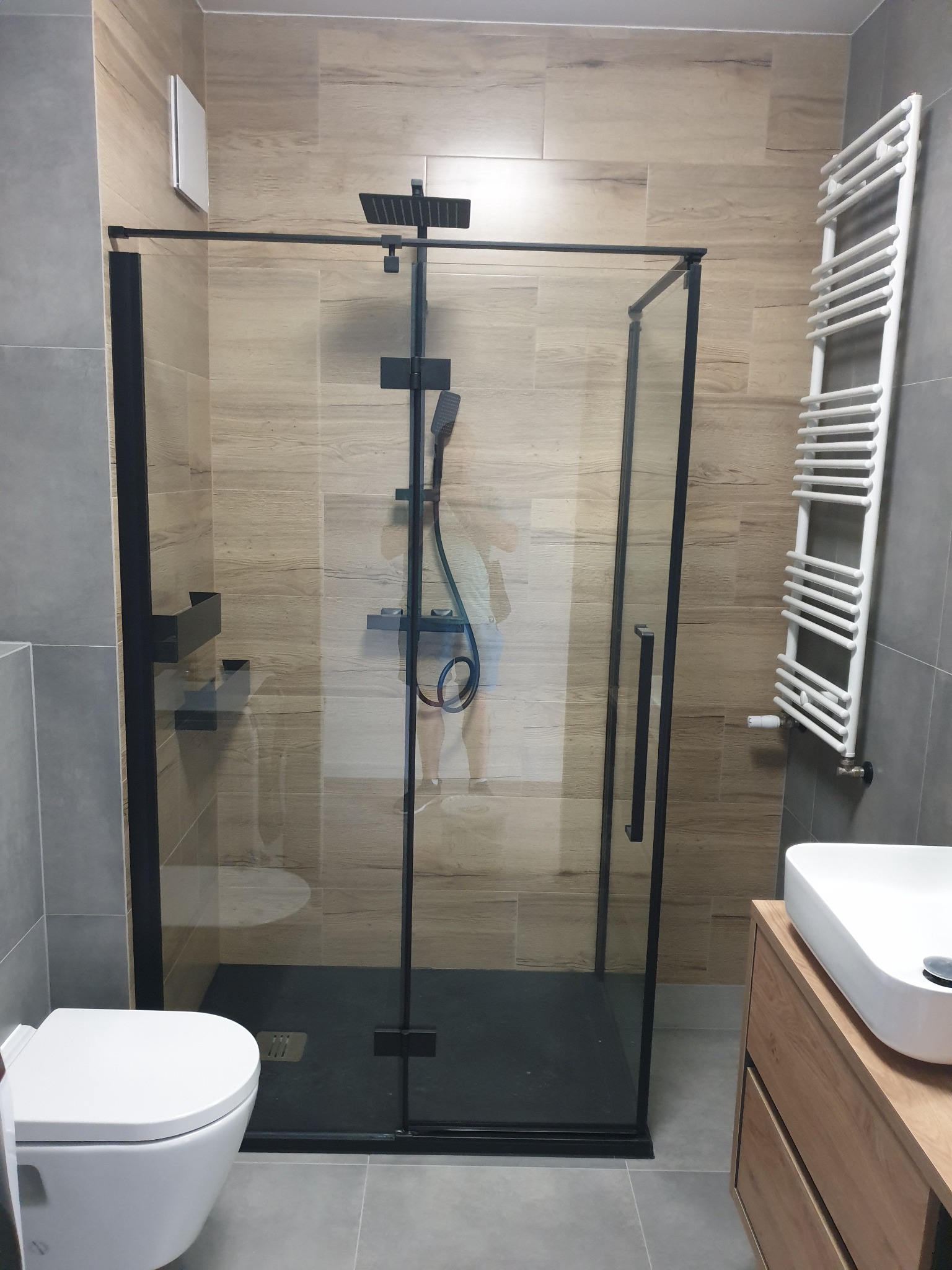 Nowoczesna kabina prysznicowa z czarnymi profilami i deszczownicą, obok umywalka nablatowa na drewnianej szafce, grzejnik drabinkowy i płytki imitujące drewno na ścianie.