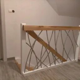 Nowoczesna balustrada schodowa z białymi elementami, drewnianą poręczą i metalowymi, zakrzywionymi prętami, widoczna na tle ściany i fragmentu schodów.