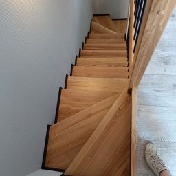 ZAKŁAD USŁUGOWY KABKEI TOMASZ NOWACKI - Kątowe ujęcie od góry na dębowe schody z czarnymi krawędziami i metalową balustradą, widoczny fragment buta na podłodze imitującej drewno.