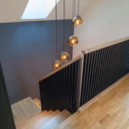 Nowoczesne schody z jasnego drewna z czarną balustradą z pionowych szczebli, oświetlone przez wiszące lampy kuliste i okno dachowe.