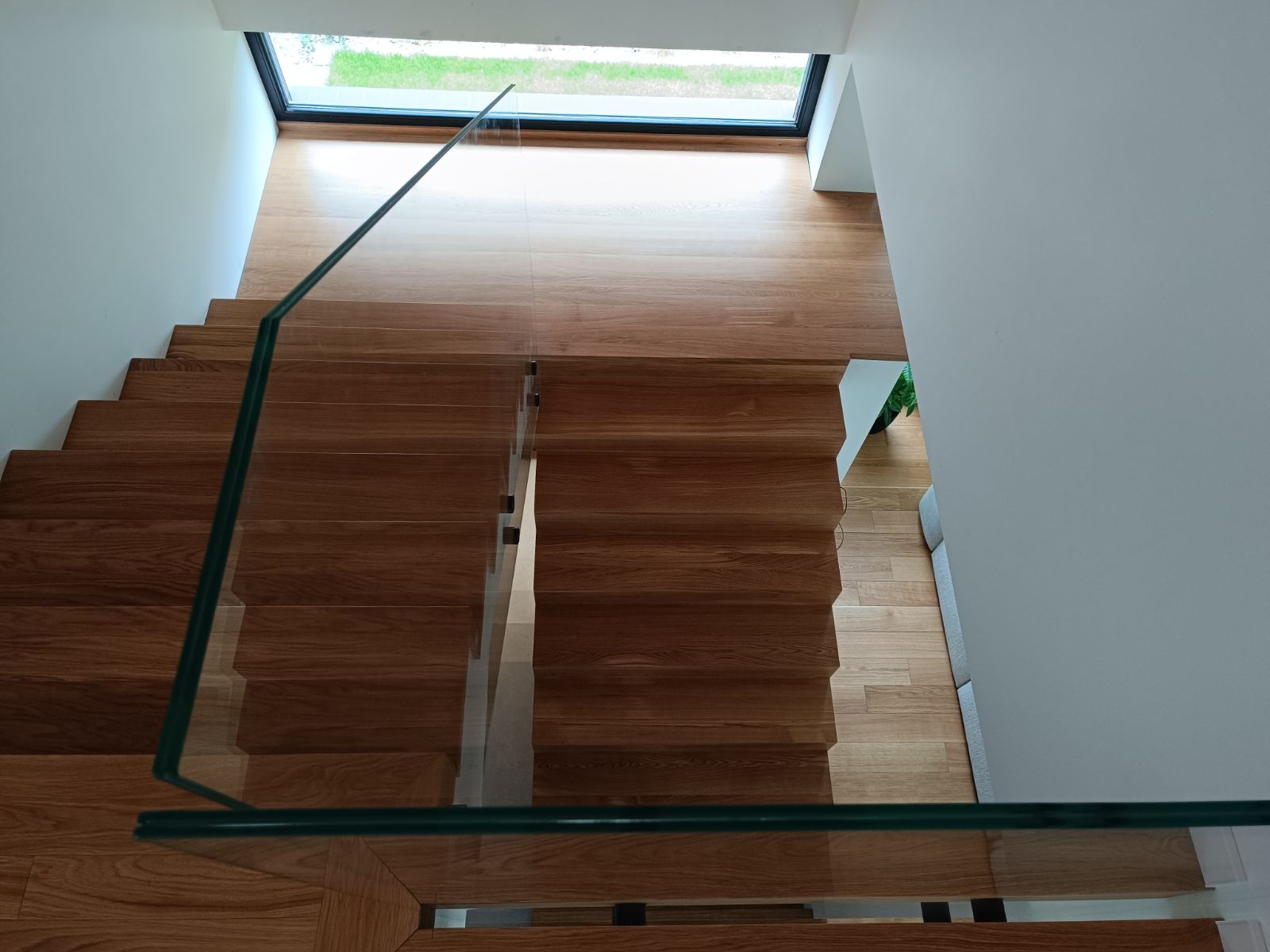 Widok z góry na drewniane schody z szklaną balustradą, prowadzące do okna dachowego z widokiem na zieleń, nowoczesne wnętrze z jasnymi ścianami.