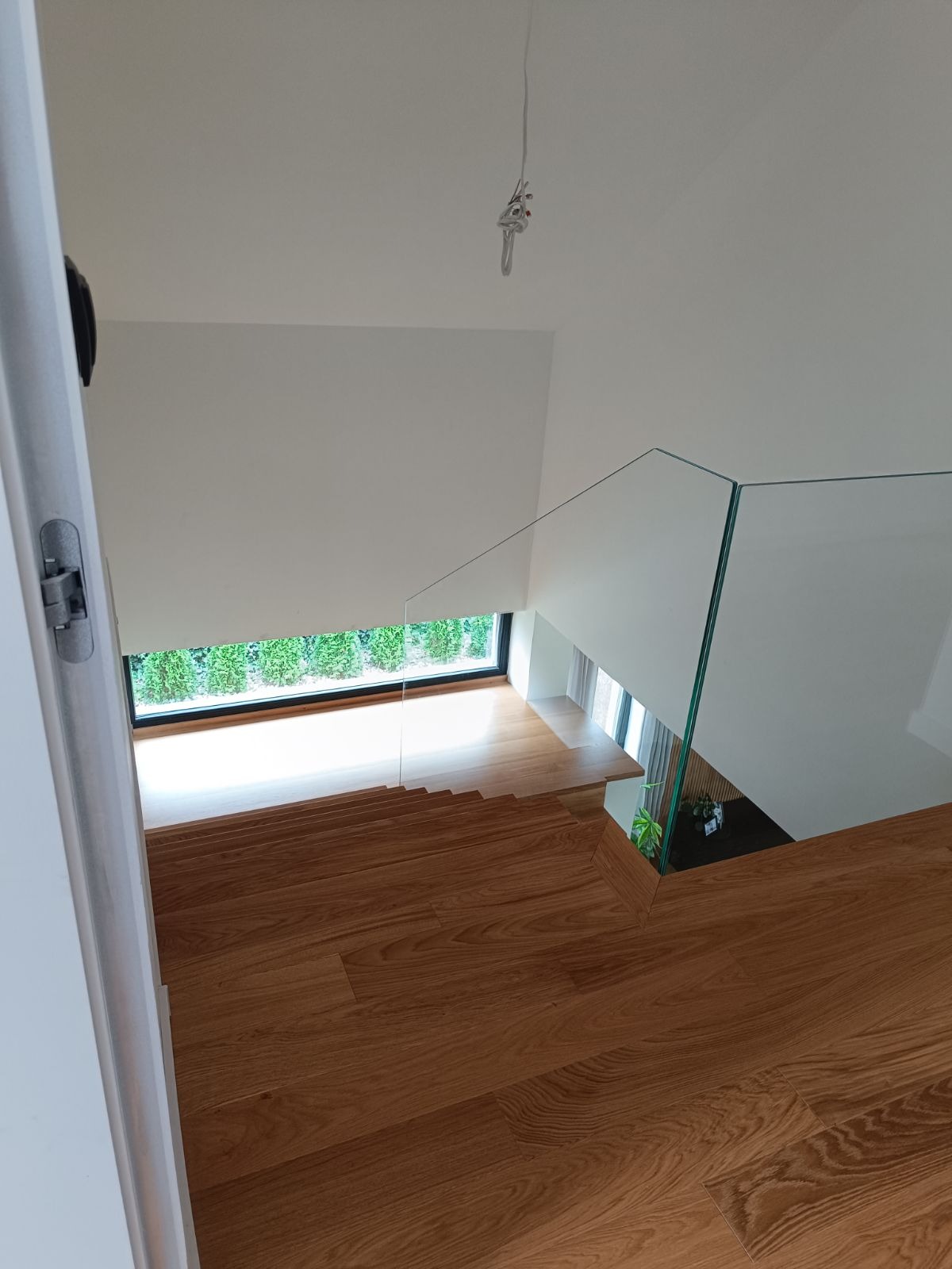 Nowoczesne schody z drewna z przeszkloną balustradą, widok z góry na jasne wnętrze z dużym oknem.