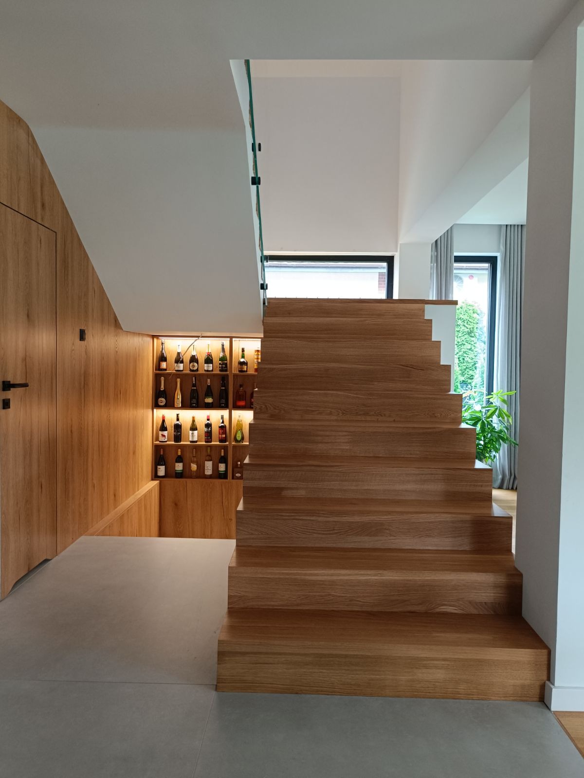 Nowoczesne, drewniane stopnie bez podstopnic, z minimalistyczną szklaną barierką, w tle podświetlana wnęka z kolekcją win.