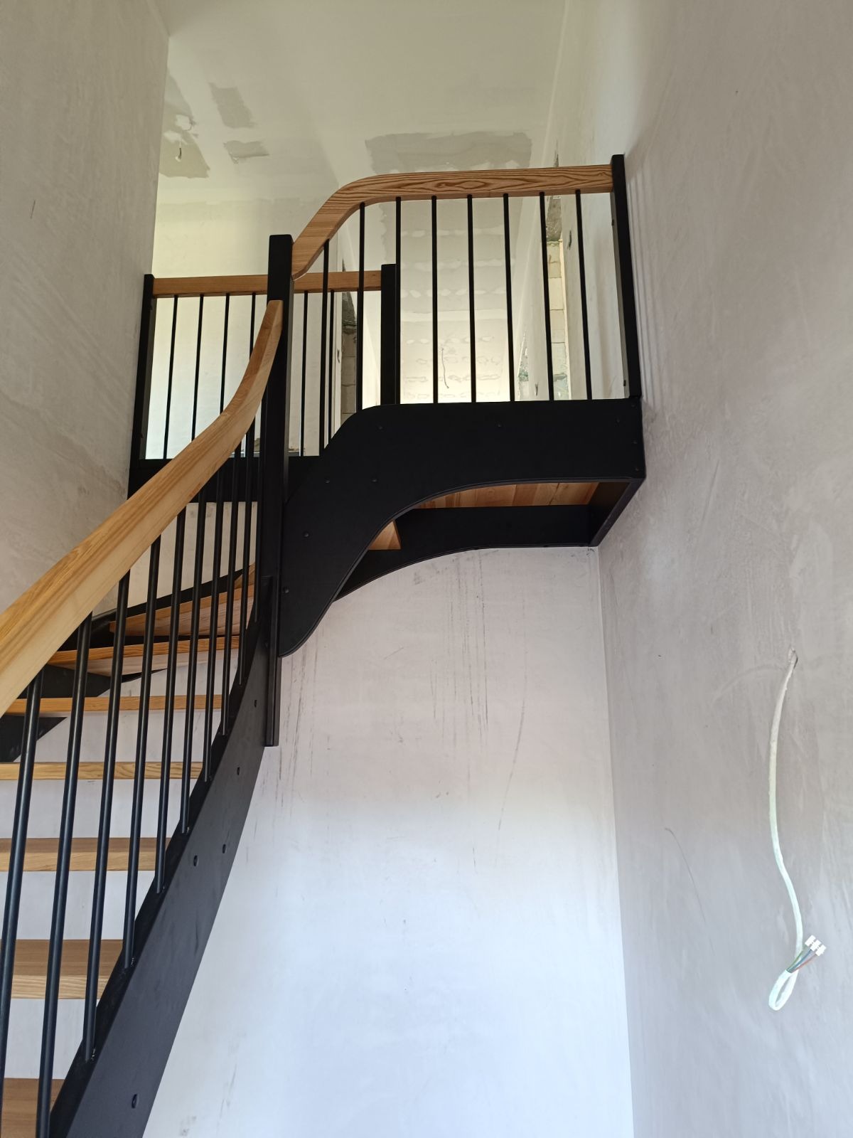 Wewnętrzne schody z drewnianymi stopniami i jasną, giętą poręczą, z czarnymi metalowymi balustradami na tle surowych ścian w trakcie wykończenia.