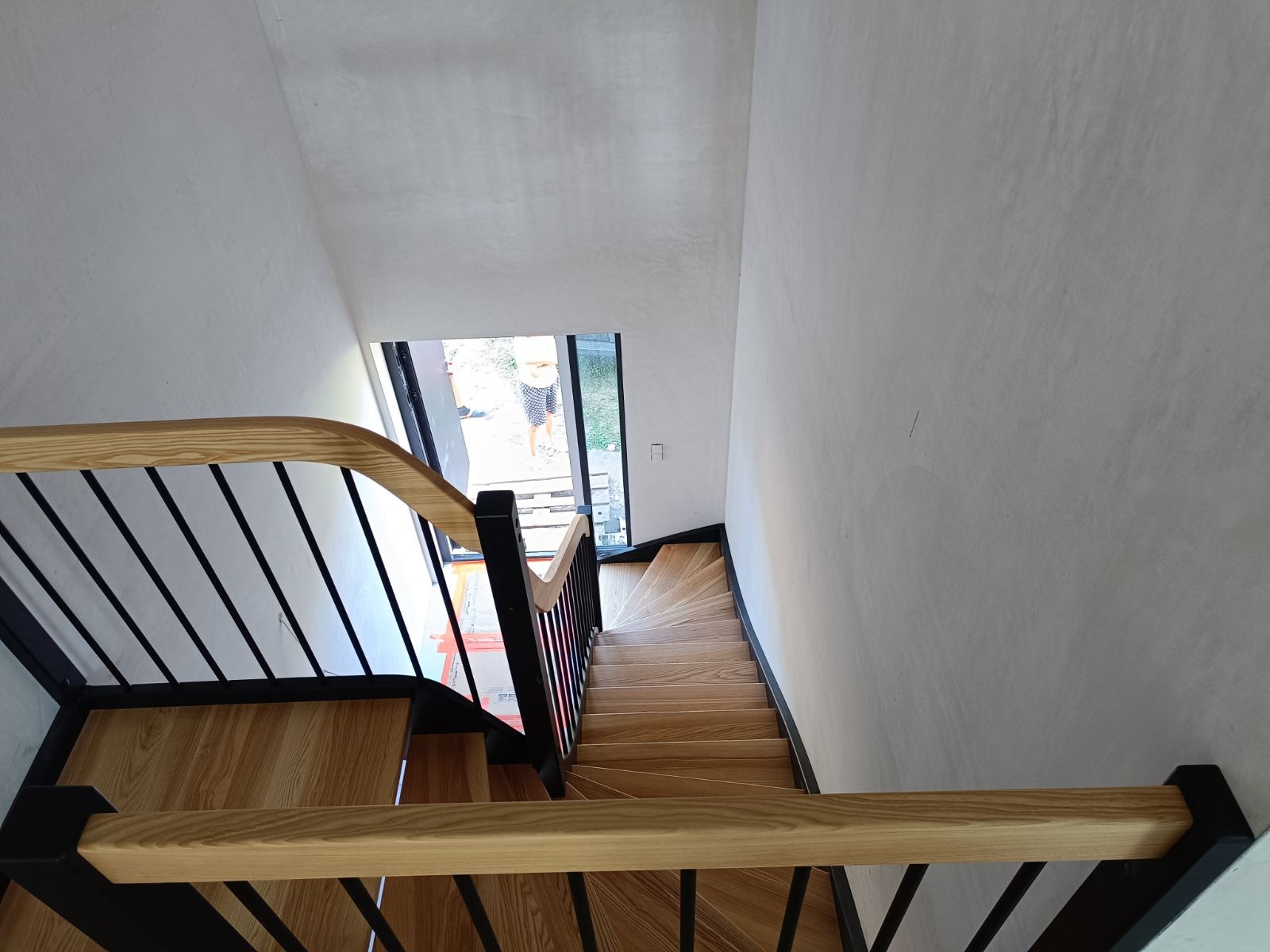 Widok z góry na drewniane schody z jasnego drewna z czarnymi metalowymi balustradami, prowadzące w dół, z oknem widocznym na końcu biegu schodów.