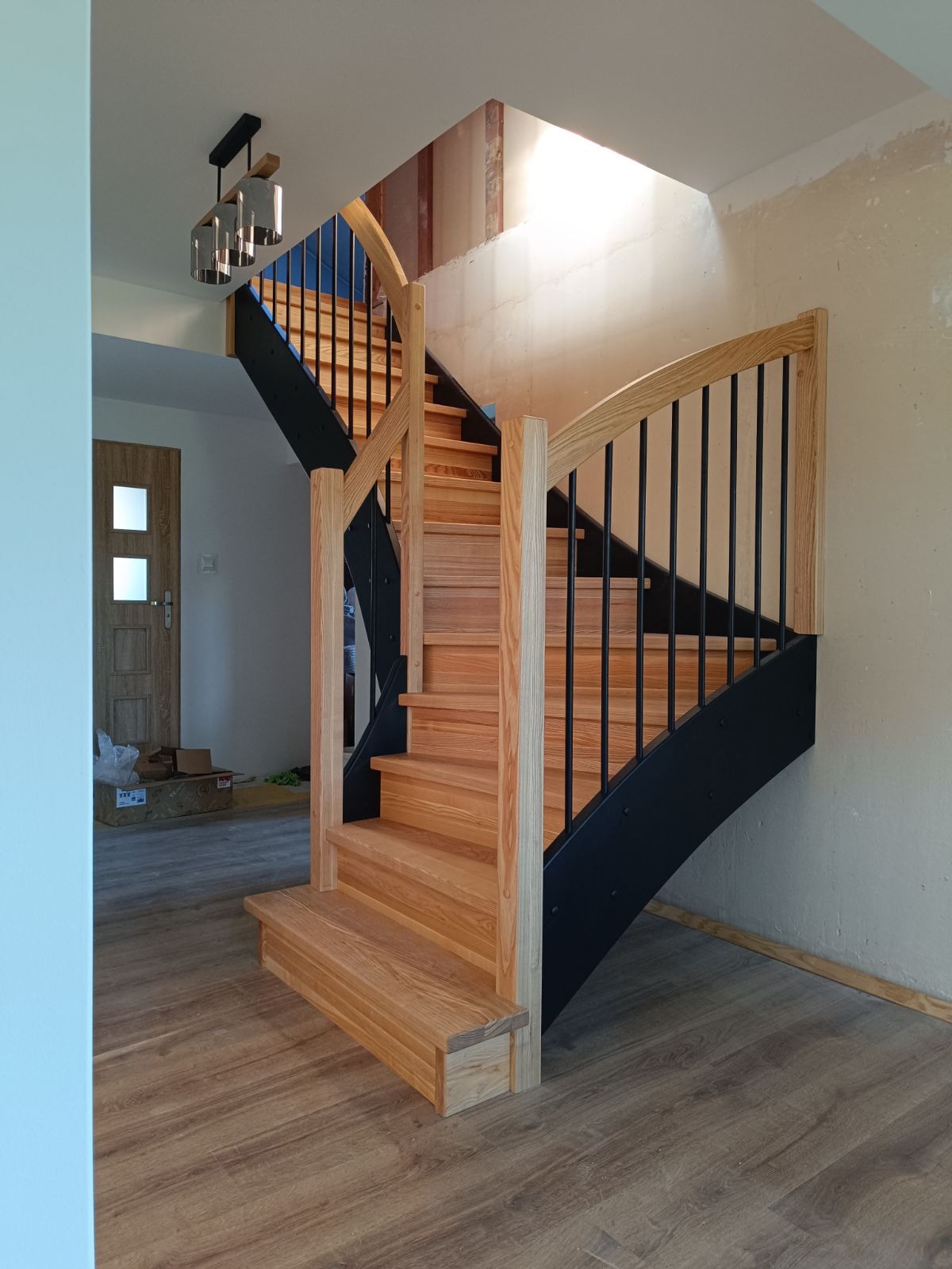 Jasne drewniane schody z czarnymi metalowymi balustradami w nowoczesnym domu, widoczne niedokończone wykończenia ścian i sufitu.