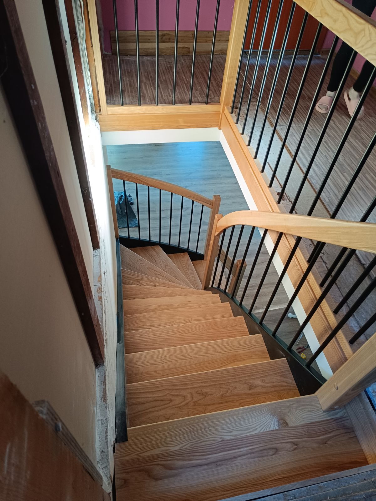 Klatka schodowa z drewnianymi schodami i czarnymi metalowymi balustradami, widok z góry na zakręcające schody, częściowo wykończone ściany i podłogi.