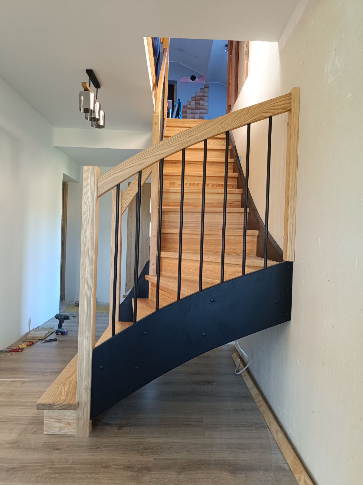 Nowoczesne schody z jasnego drewna z czarnymi metalowymi balustradami, widoczne narzędzia na podłodze, wnętrze w trakcie wykończenia.