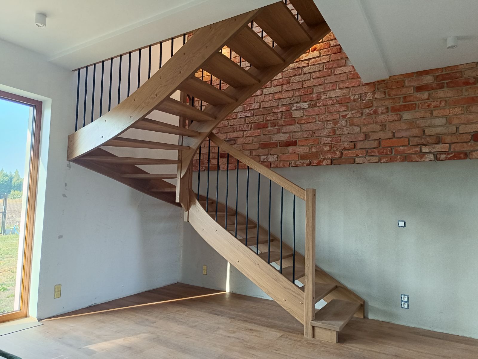 Jasne, drewniane schody z czarnymi, metalowymi balustradami, zakręcające w lewo, z widoczną ścianą z czerwonej cegły w tle, oświetlone naturalnym światłem wpadającym przez okno.
