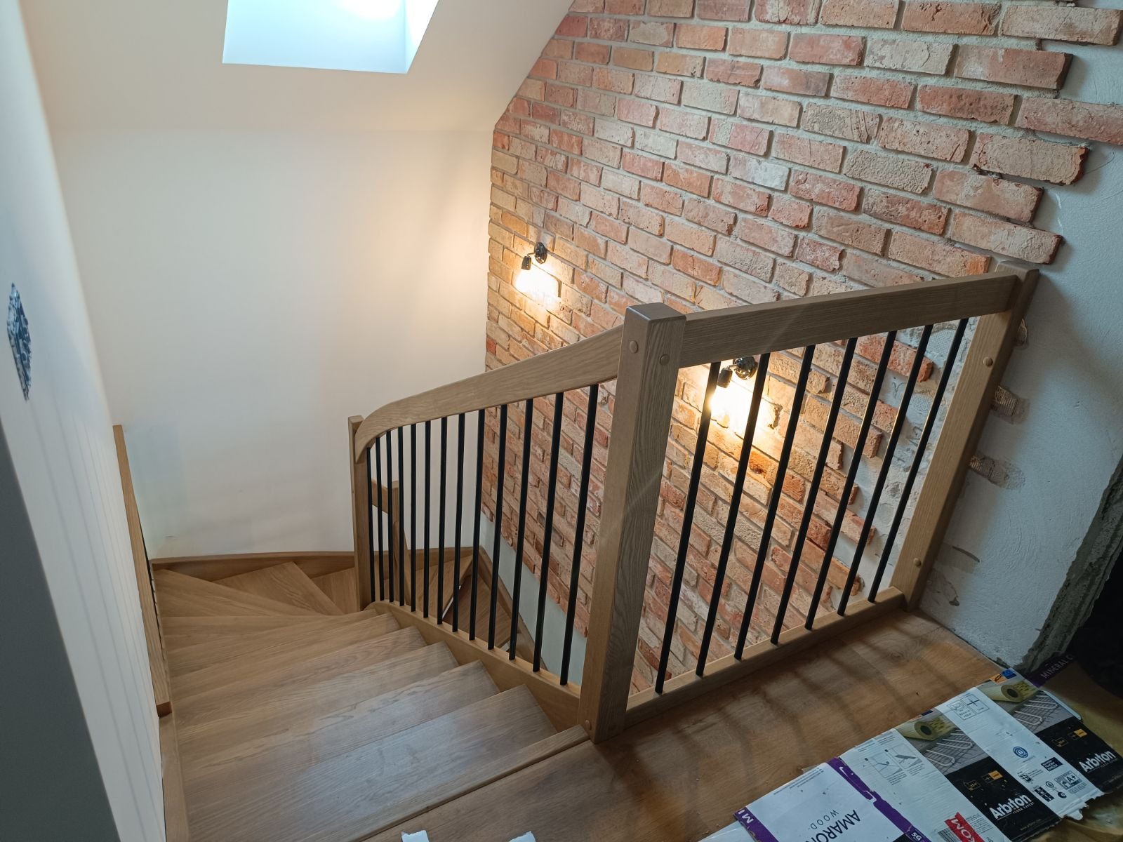 Jasne drewniane stopnie schodów z czarnymi metalowymi prętami balustrady i drewnianą poręczą, oświetlone lampami na ścianie z cegły i światłem z okna dachowego.