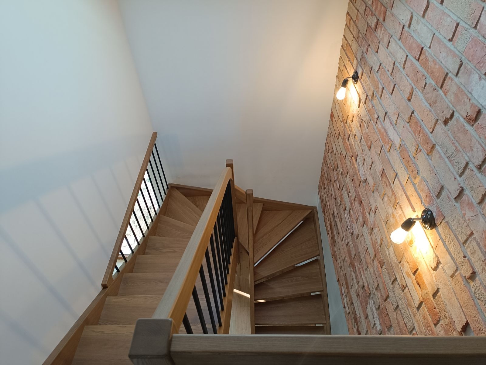 Jasne, drewniane schody z czarnymi, metalowymi balustradami, widok z dołu, ściana z cegły z dwoma żarówkami w tle.