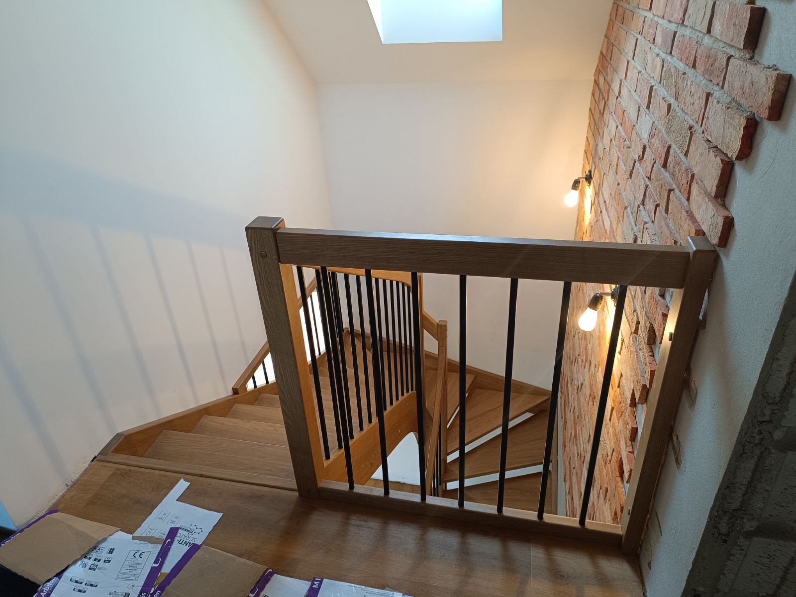 Widok z góry na drewniane schody z czarnymi, metalowymi szprosami w balustradzie, oświetlone żarówkami na ceglanej ścianie i światłem z okna dachowego.