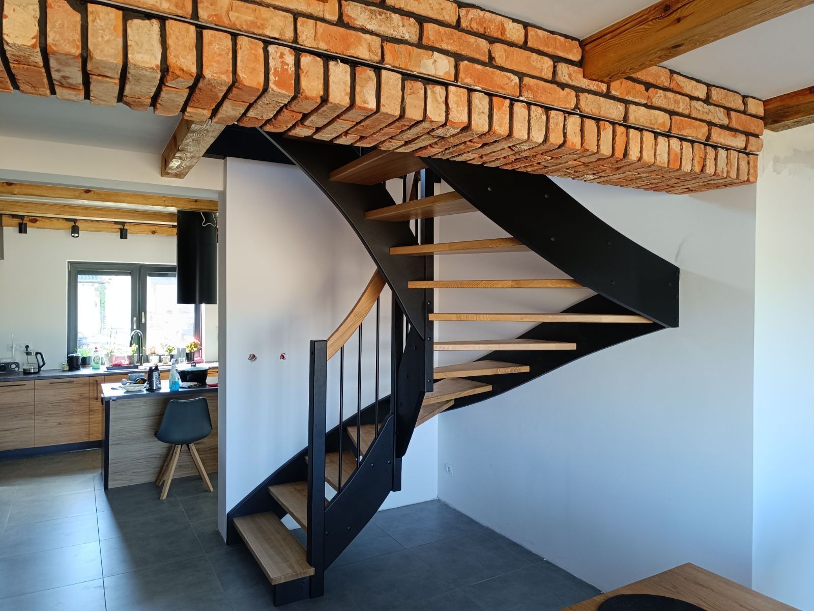 Nowoczesne schody drewniane z czarną metalową konstrukcją w jasnym wnętrzu z ceglanym sufitem, widok na kuchnię w tle.