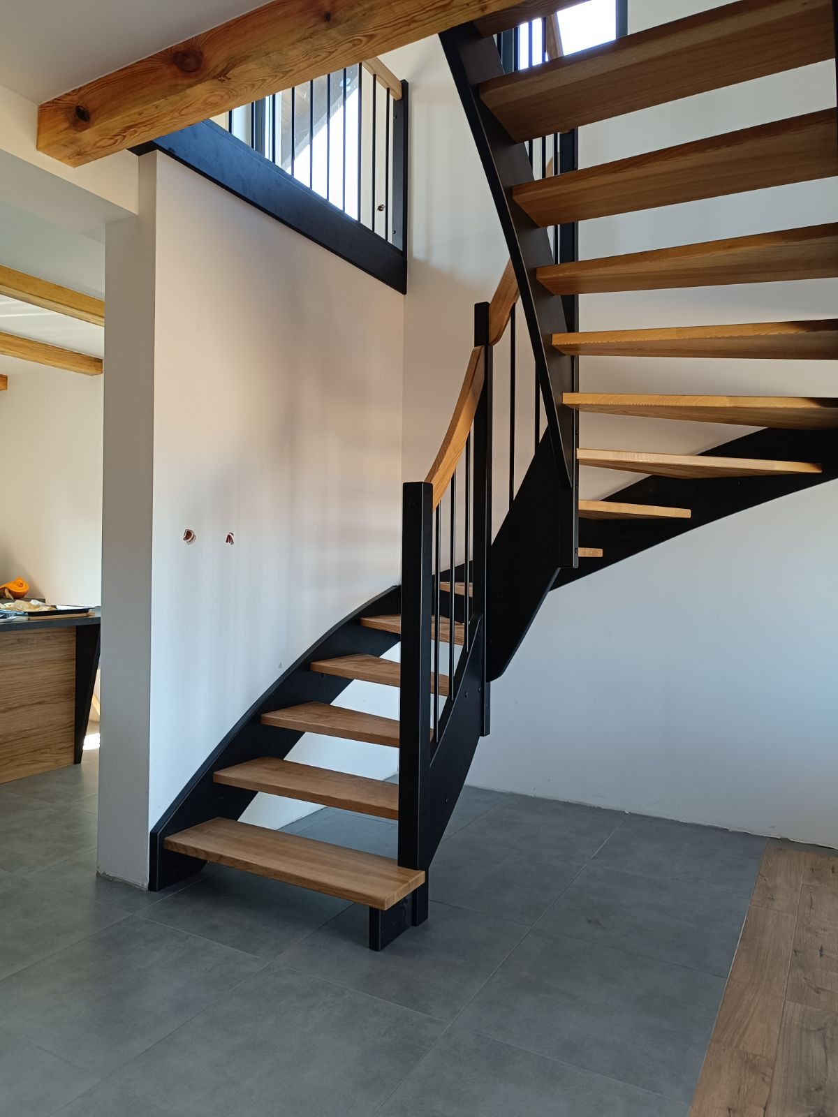 Spiralne schody drewniane z czarną metalową konstrukcją i drewnianą balustradą, widok z dołu, szare płytki podłogowe, fragment salonu w tle.