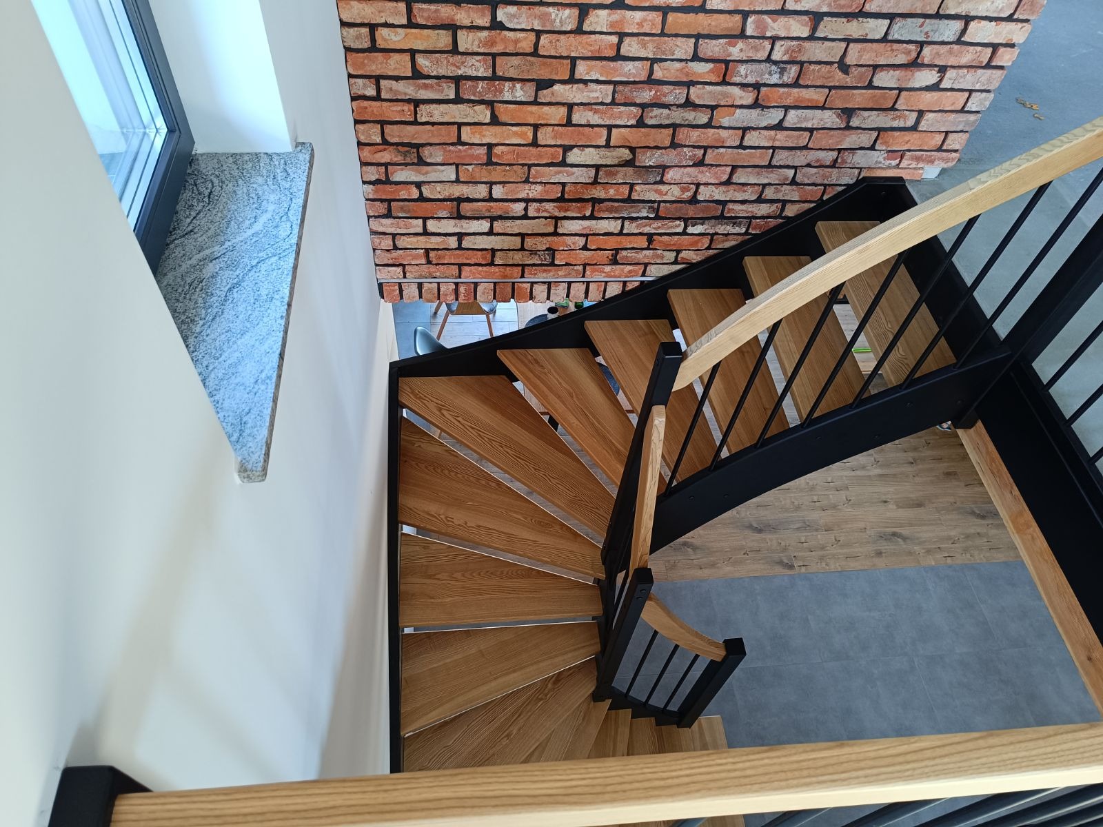 Spiralne schody z jasnego drewna, z czarnymi metalowymi elementami konstrukcyjnymi i drewnianą balustradą, widok z góry, ściana wykończona cegłą.