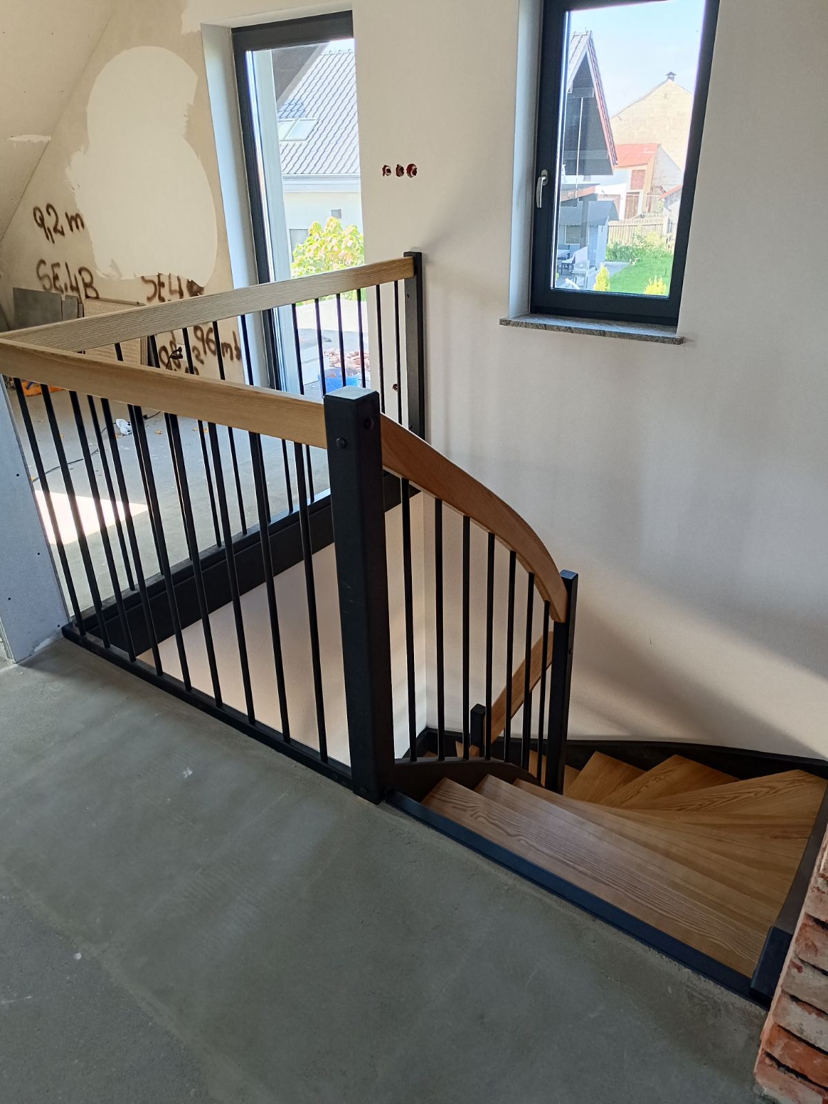 Kręte schody z jasnego drewna z czarnymi metalowymi balustradami w surowym wnętrzu, widok z góry na dół, ściany z widocznymi śladami prac wykończeniowych, okno z widokiem na domy w tle.