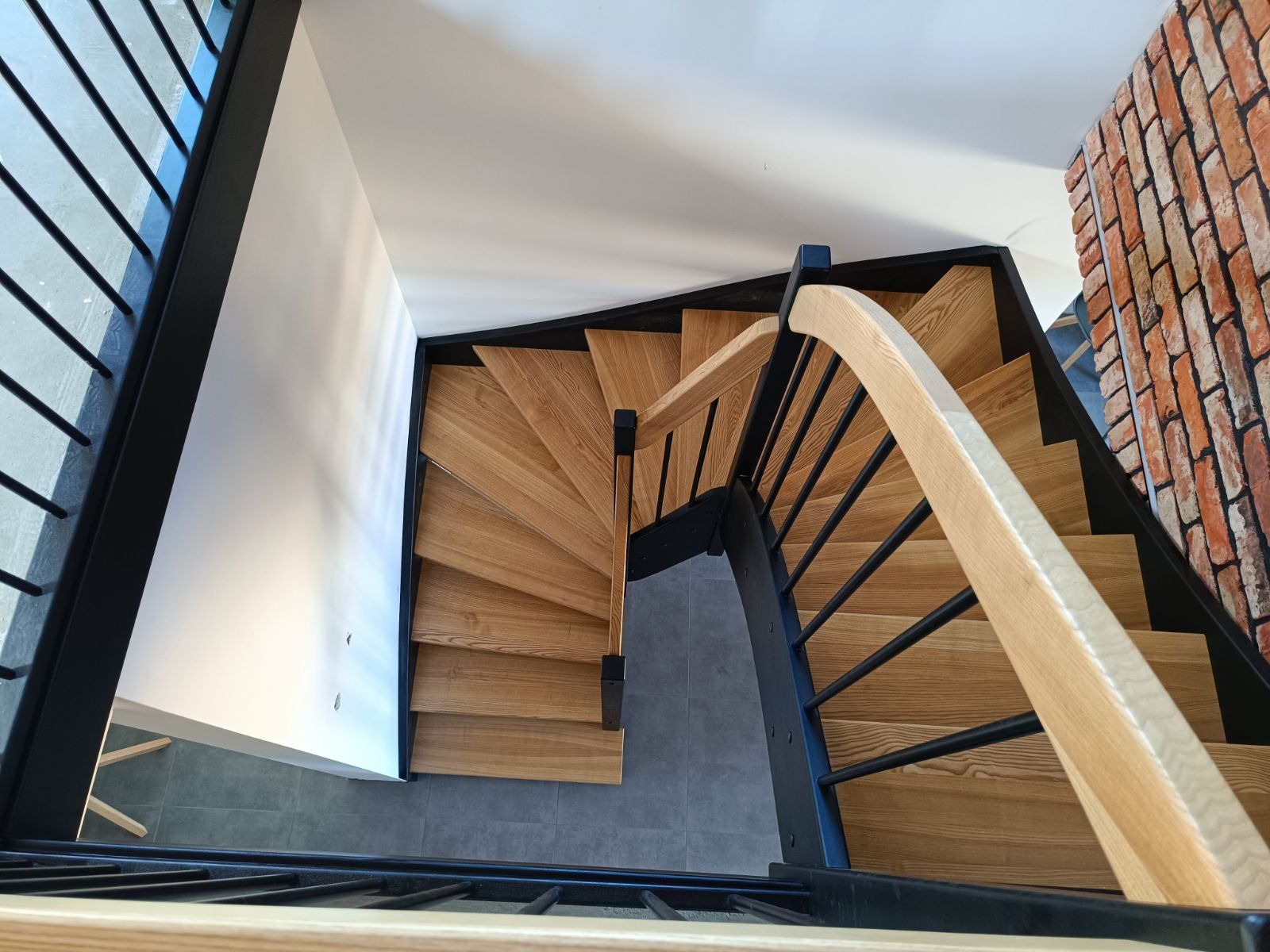 Widok z góry na spiralne schody z jasnego drewna z czarną metalową konstrukcją i jasną poręczą, ściana z cegły w tle.