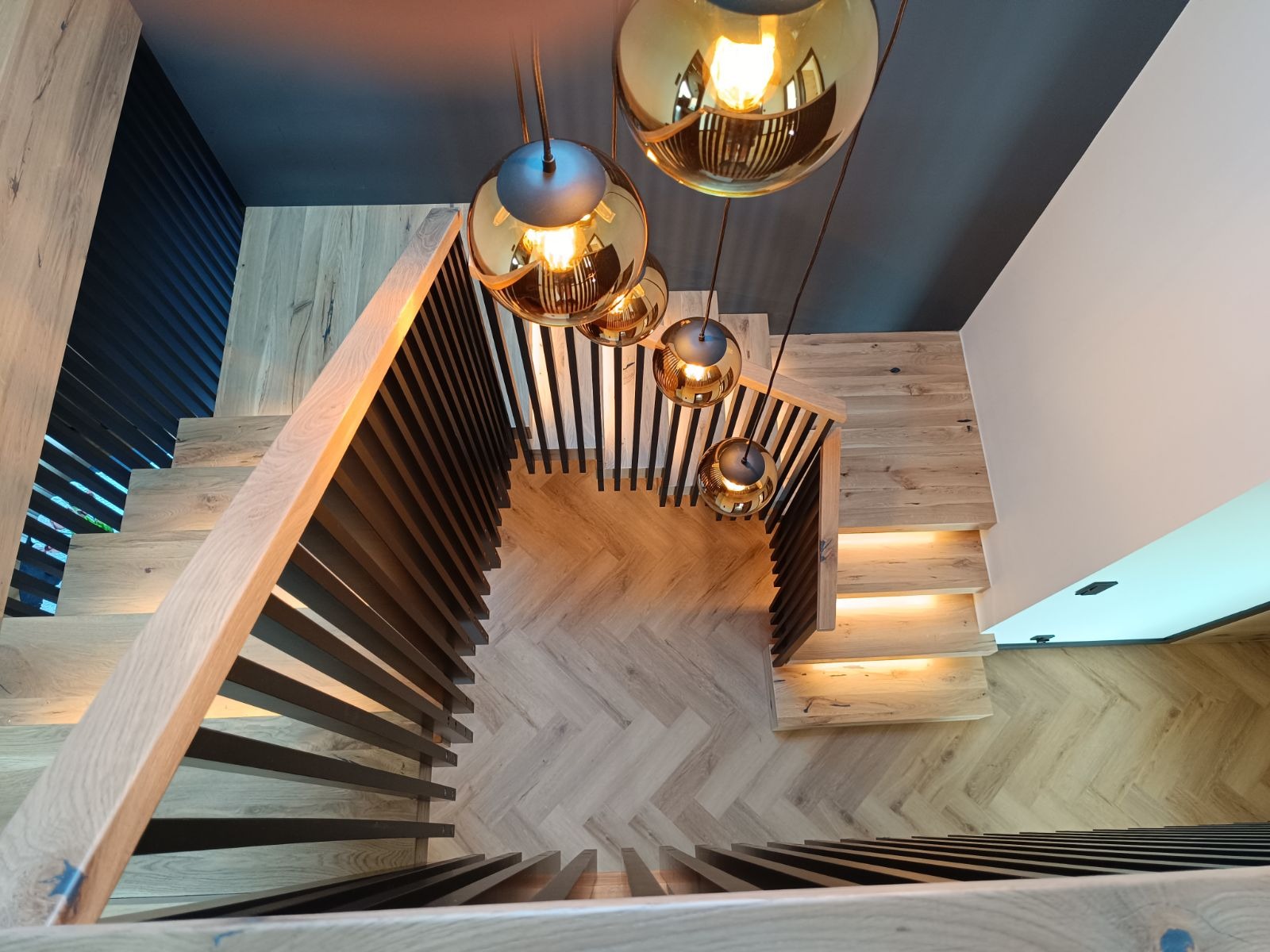 Widok z góry na nowoczesne schody z jasnego drewna z podświetleniem, z ciemnymi pionowymi balustradami, uzupełnione wiszącymi lampami o złotej barwie, na tle parkietu ułożonego w jodełkę.