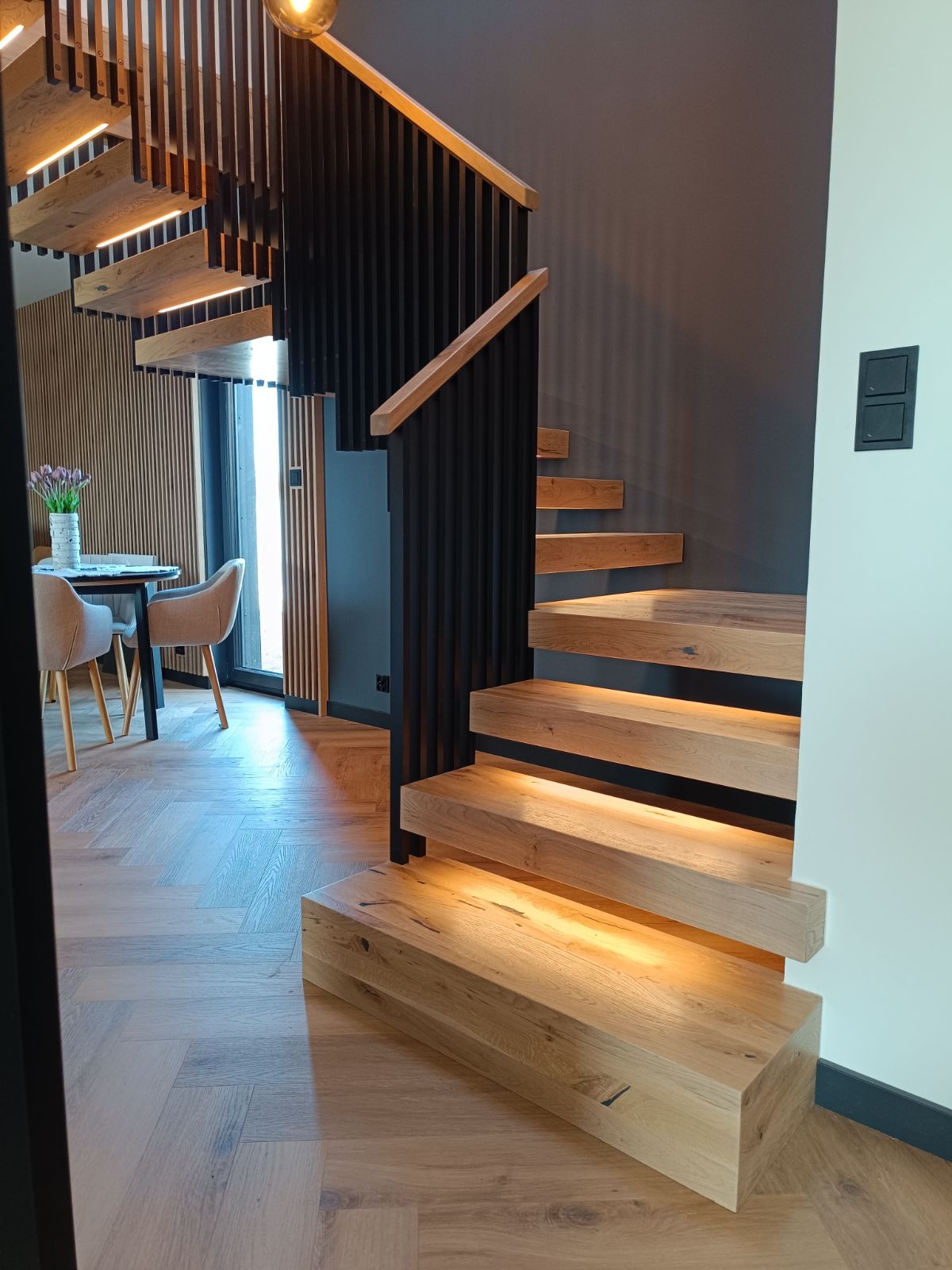 Nowoczesne schody z podświetlanymi stopniami, drewniane stopnie, czarna metalowa balustrada, widok z dołu na salon z drewnianą ścianą i stołem.