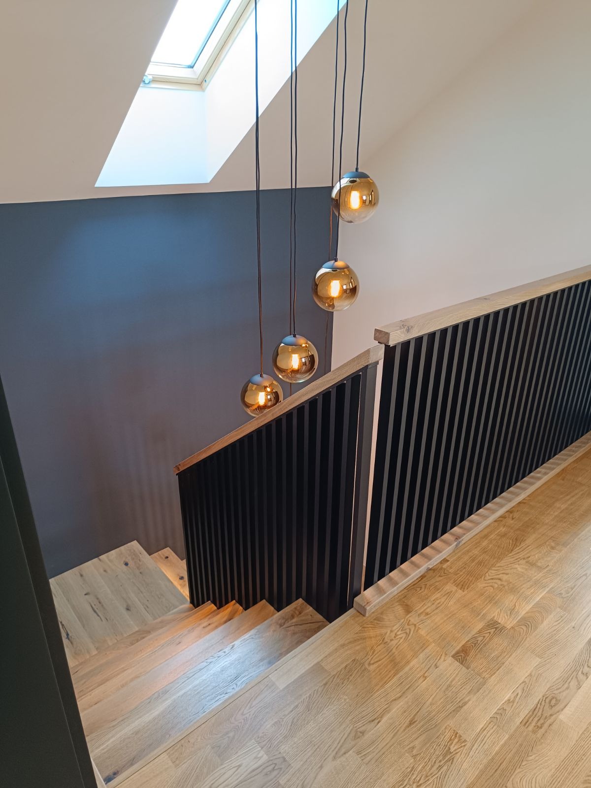 Nowoczesne schody z jasnego drewna z czarną balustradą z pionowych szczebli, oświetlone przez wiszące lampy kuliste i okno dachowe.