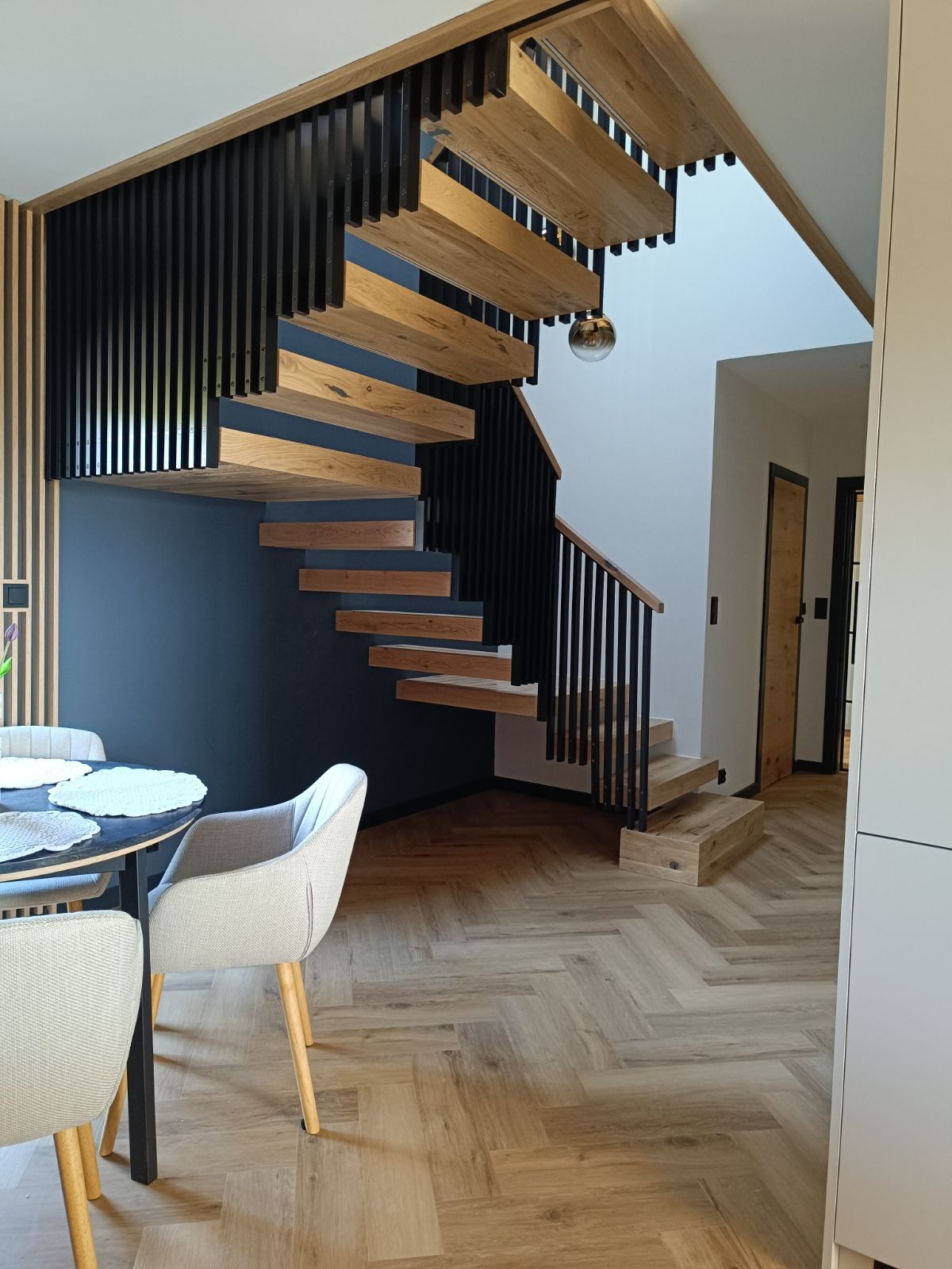 Nowoczesne, wiszące stopnie z drewna dębowego z czarną, pionową balustradą, zamontowane przy ciemnej ścianie, widoczne z perspektywy jadalni z parkietem w jodełkę.
