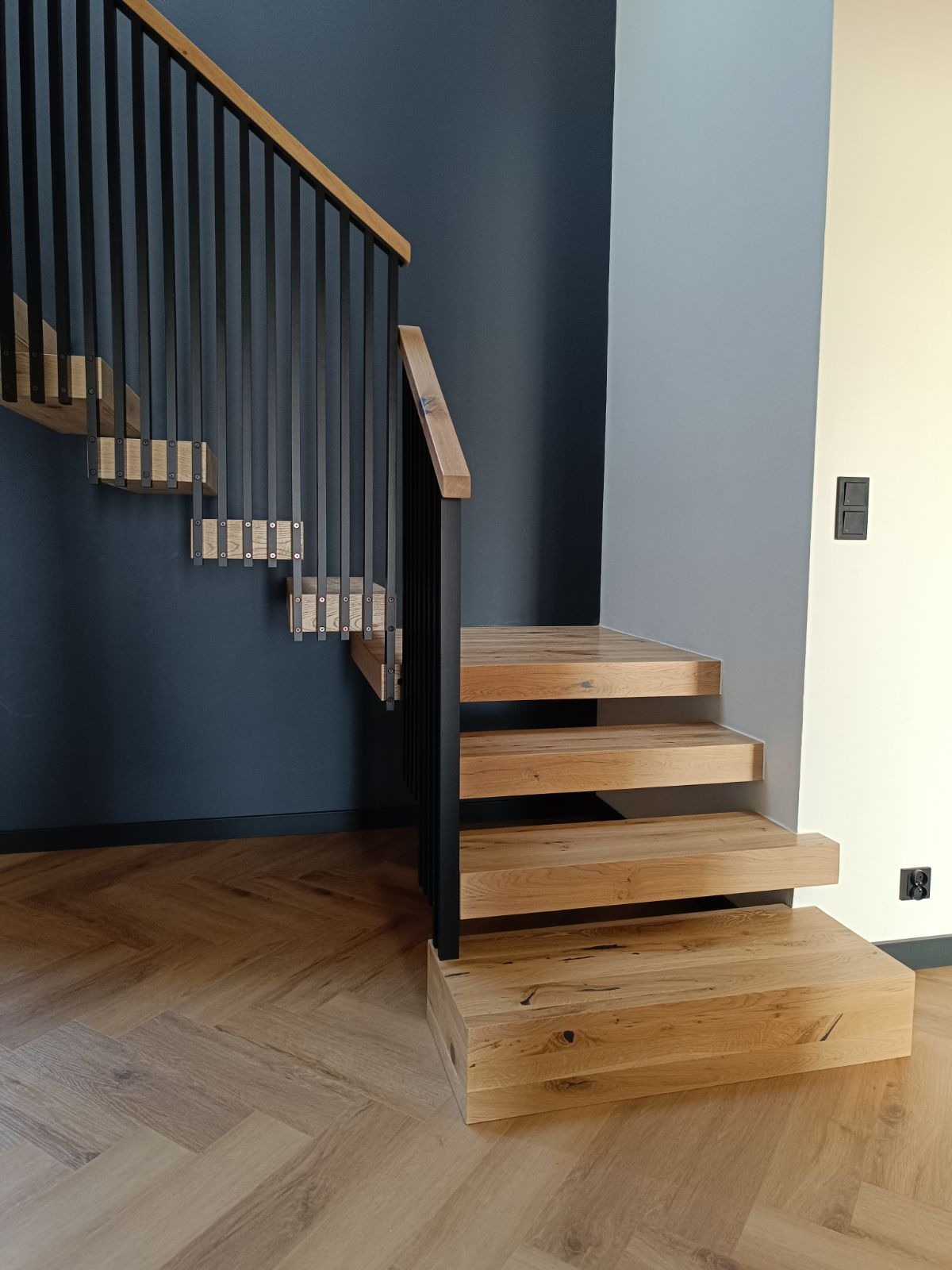 Nowoczesne, dębowe stopnie schodowe wspornikowe z czarną balustradą na tle ściany w odcieniach szarości i granatu, parkiet ułożony w jodełkę.