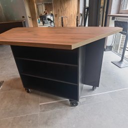 Fabryka mebli na wymiar - Mobilny barek z blatem drewnianym i czarną podstawą na kółkach, widoczny na tle wnętrza z drewnianymi elementami i oknem. Ujęcie z niskiego kąta.